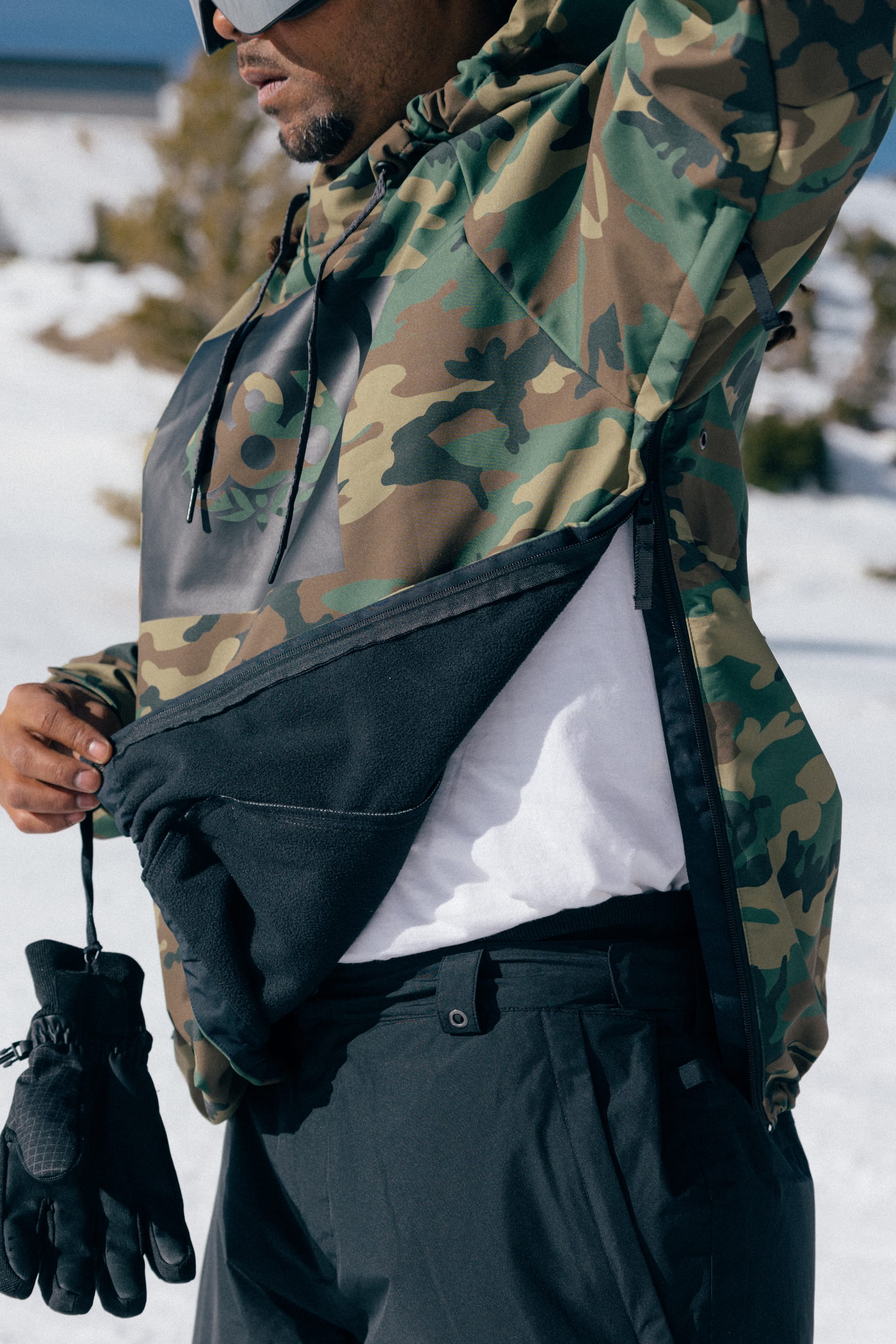 Wearing the 686 Men’s Waterproof Hoody by 686, a person stands in a snowy outdoor setting, unzipping the side to reveal a white shirt underneath and holding black gloves.