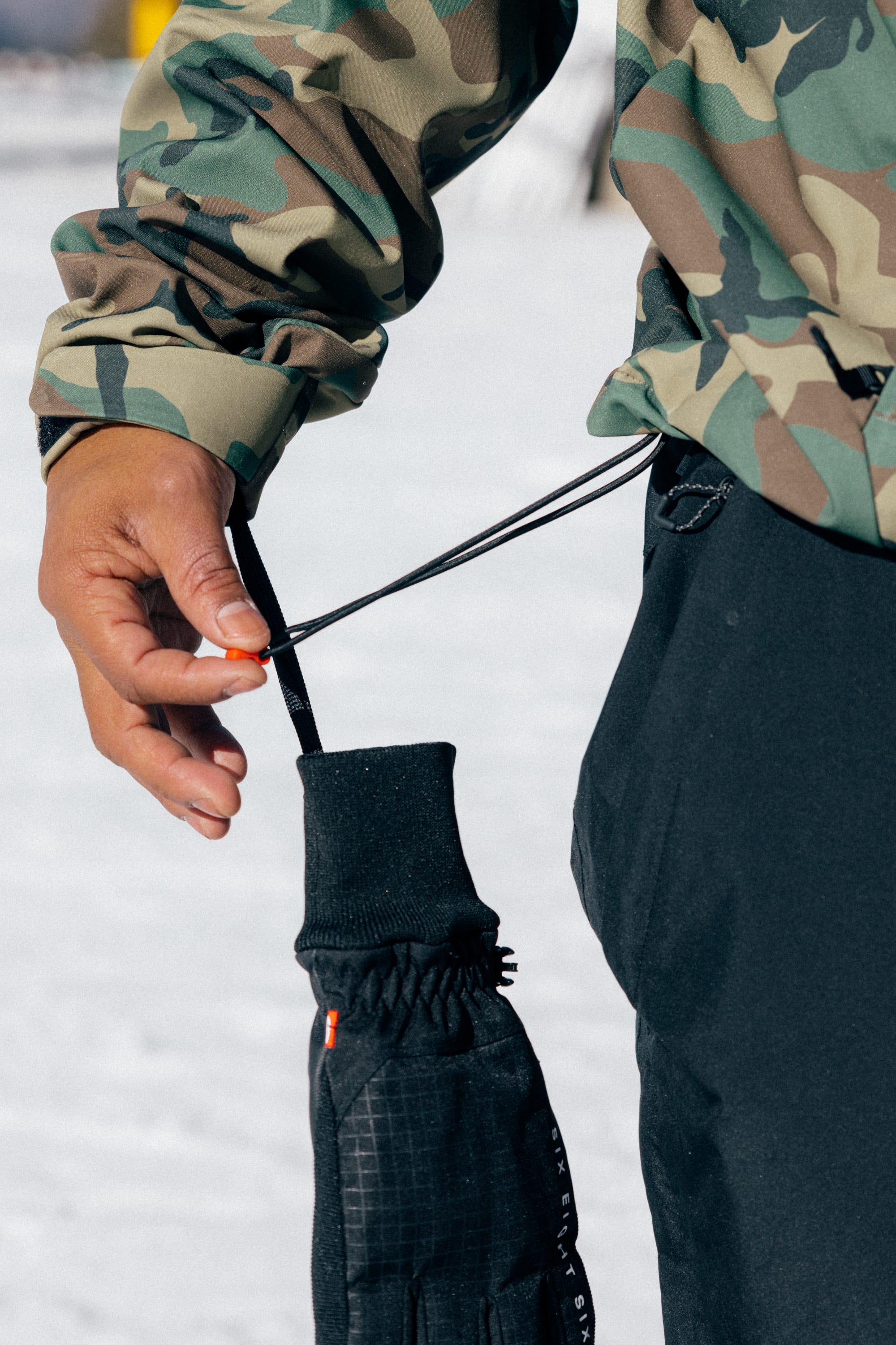 Wearing the 686 Men’s Waterproof Hoody, a person stands outdoors on a snowy surface, dressed in black pants and adjusting the wrist strap of a black glove.