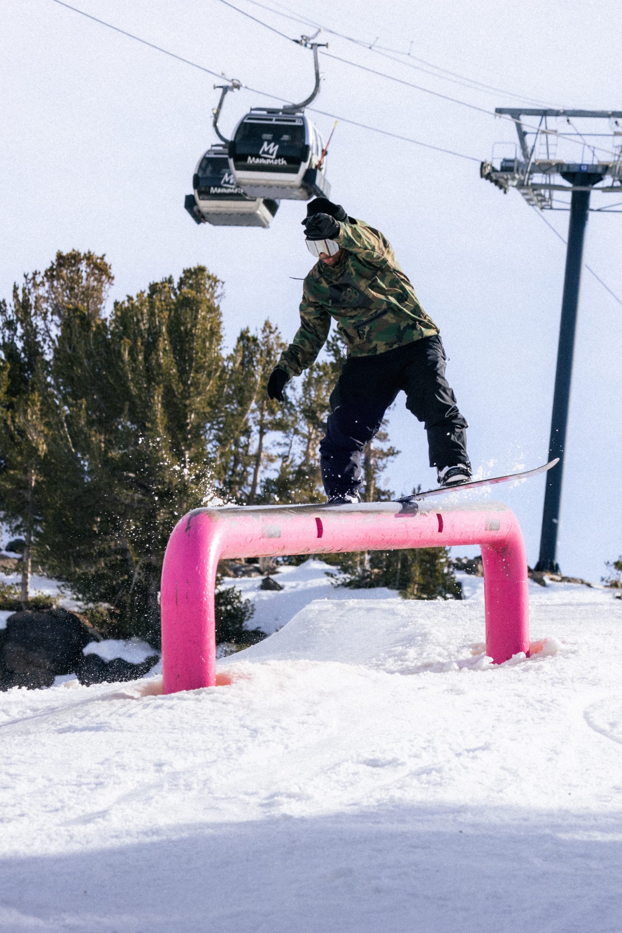 Wearing the 686 Men’s Waterproof Hoody, a snowboarder grinds on a bright pink rail in a snowy terrain park, surrounded by trees and a ski lift in the background.