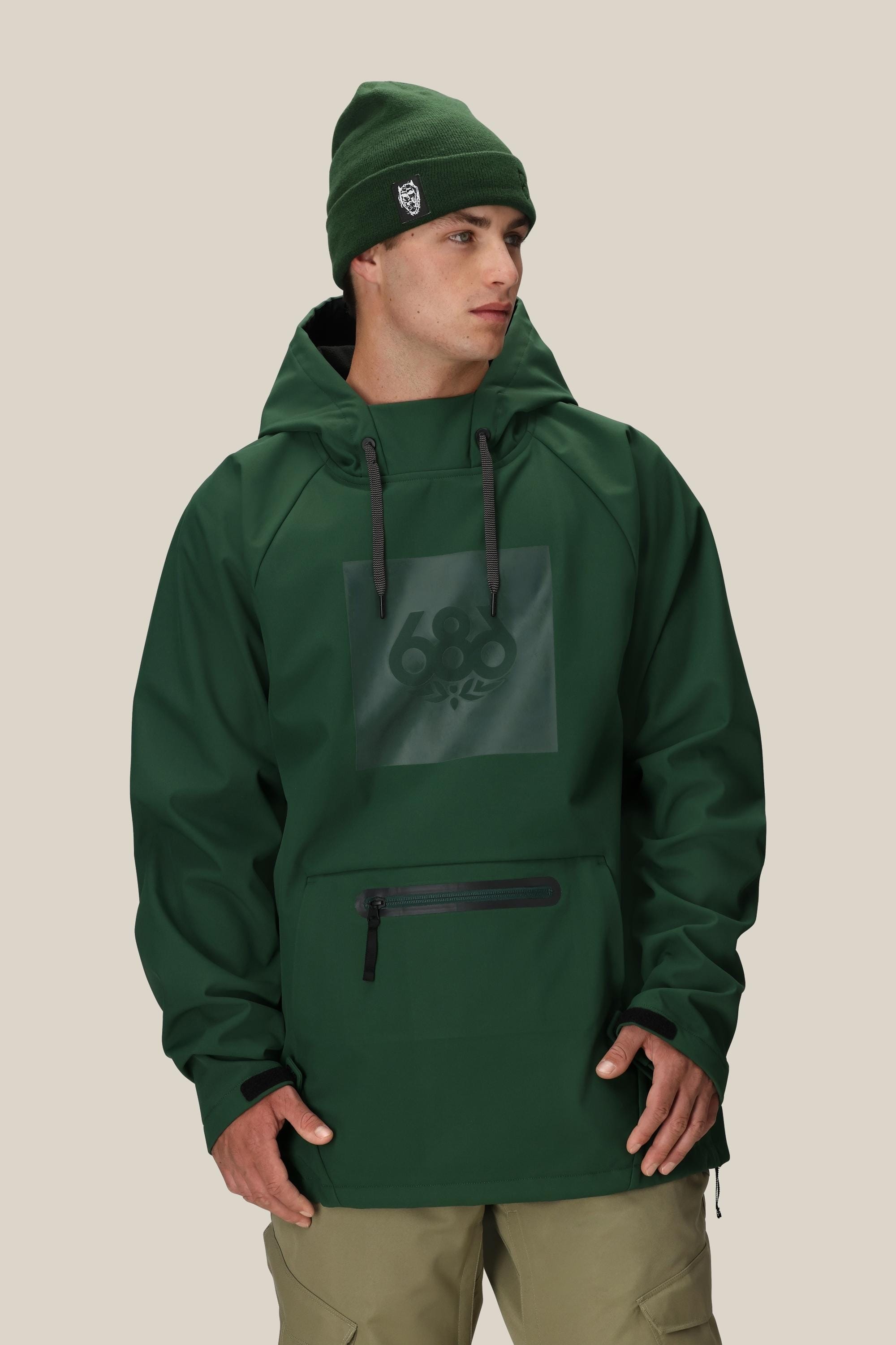 A man wearing the 686 Men’s Waterproof Hoody in dark green—with a front zipper pocket and the 686 logo on the chest—paired with khaki pants stands against a plain beige background.