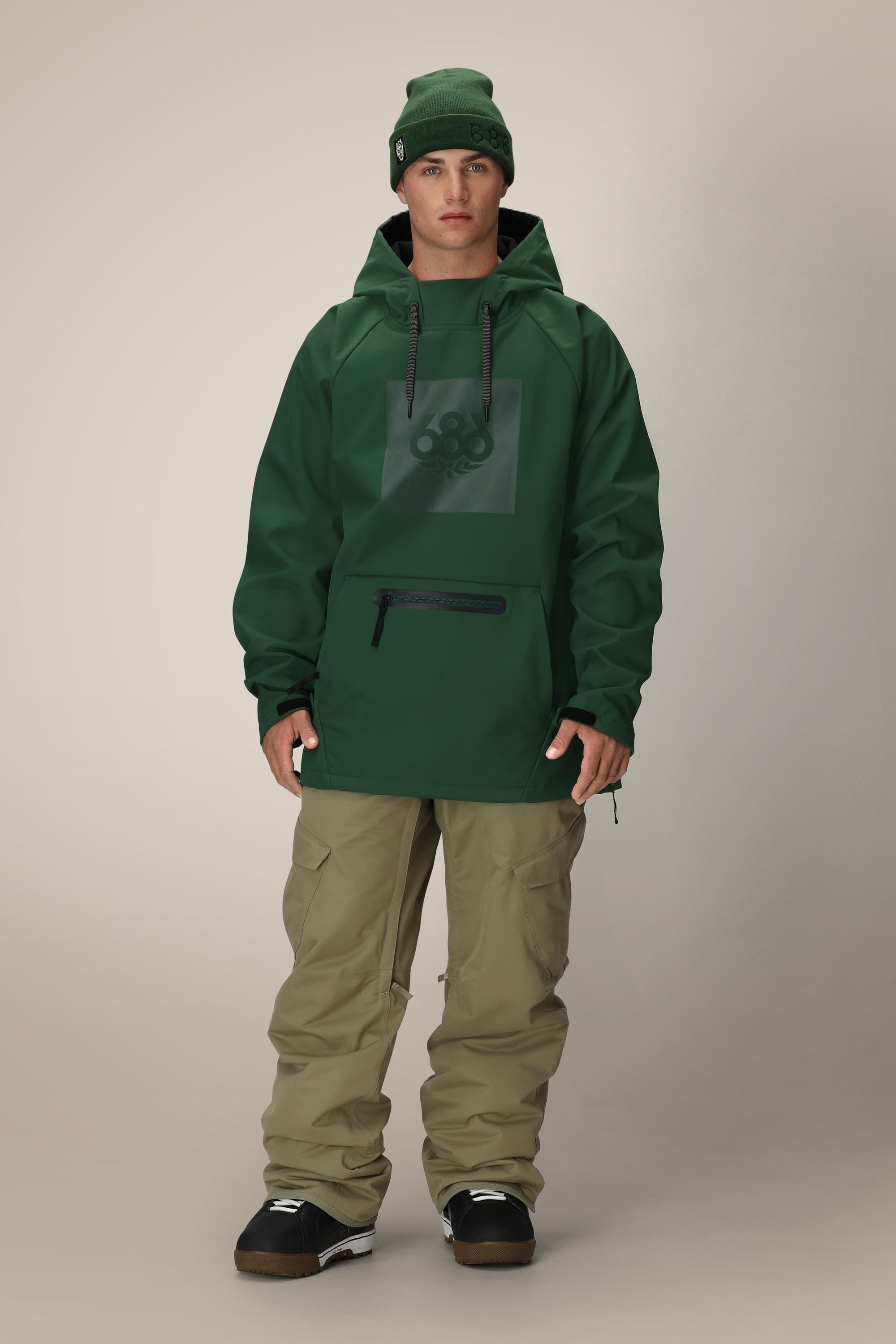 A person wearing the 686 Men’s Waterproof Hoody by 686, olive cargo pants, black snow boots, and a green beanie stands against a plain, light background.
