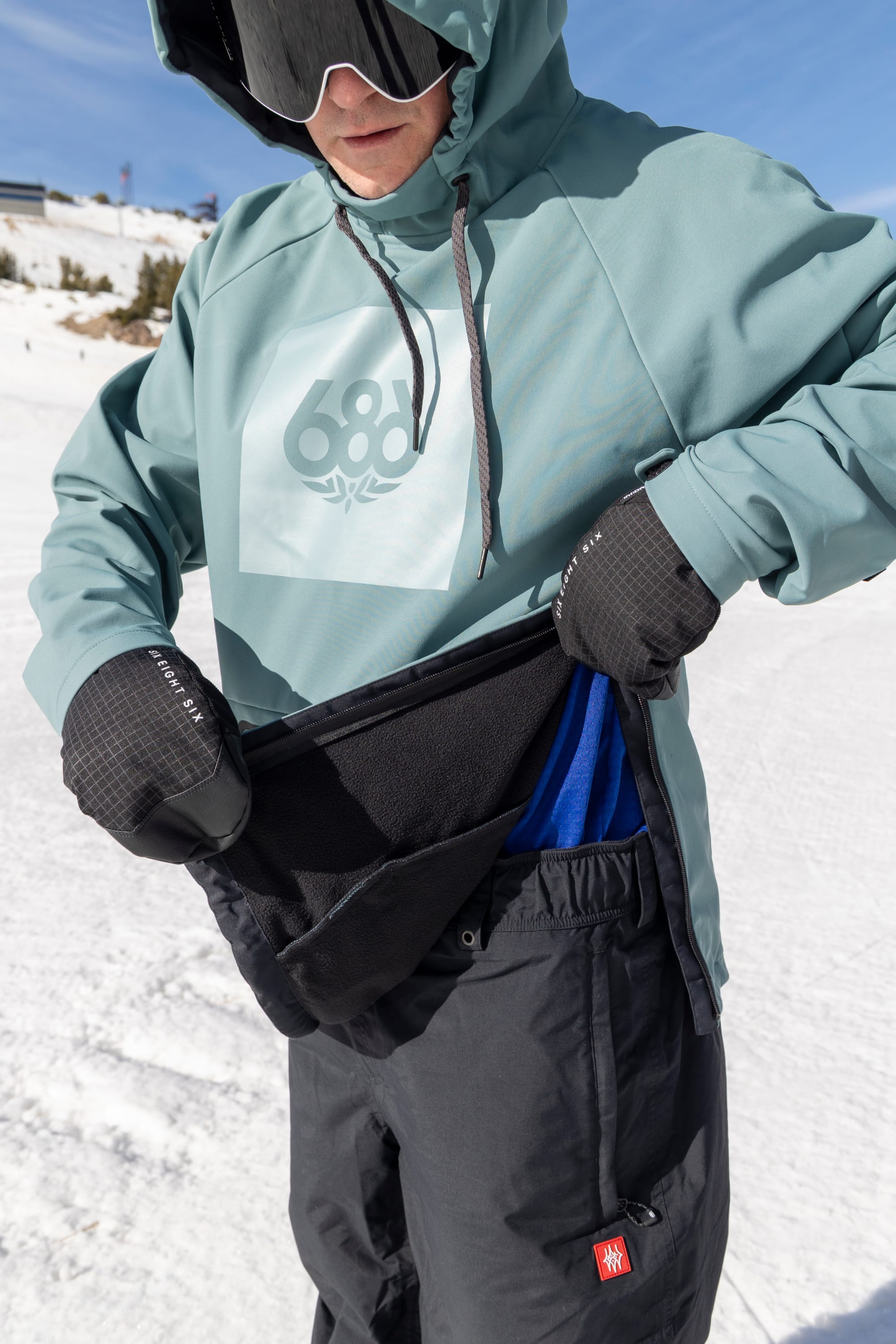 A person in winter gear pulls up their 686 Men’s Waterproof Hoody, revealing a fleece waist warmer over a blue shirt. They wear black gloves and stand on a snowy slope under a sunny sky.