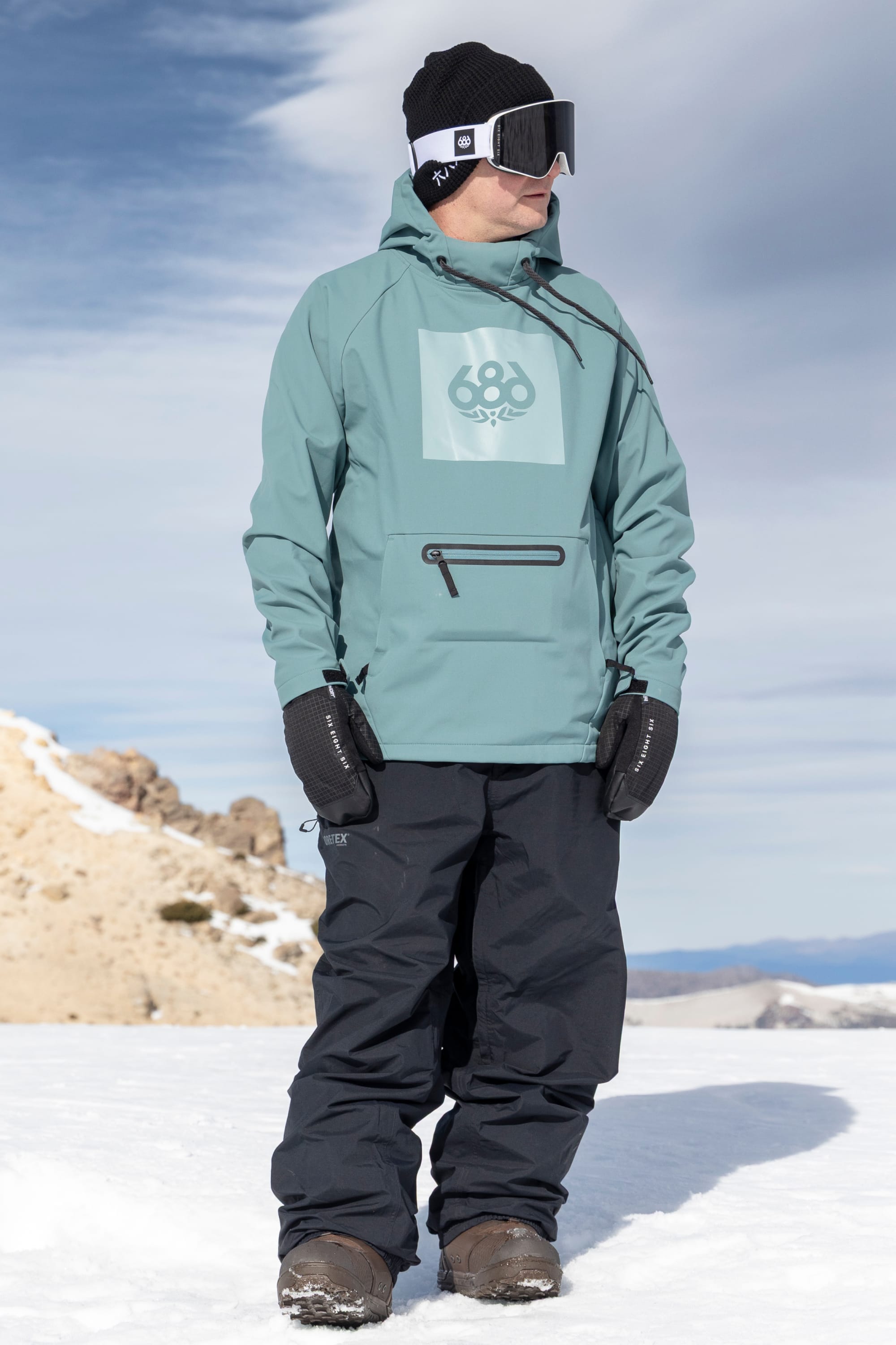 A person wearing the 686 Men’s Waterproof Hoody by 686, along with black pants, gloves, hat, and goggles, stands on a snowy landscape under a cloudy sky.