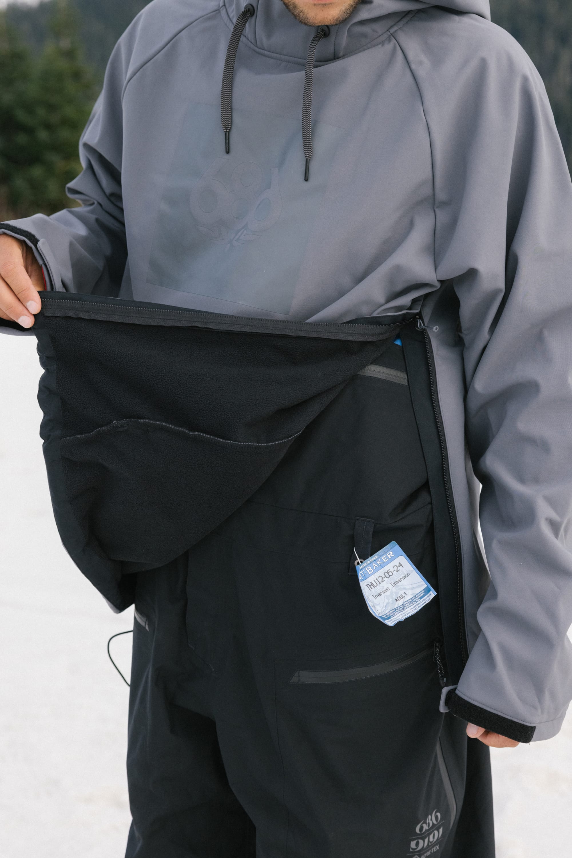A person wearing the 686 Men’s Waterproof Hoody and black snow pants lifts a flap to reveal a ski pass. Only the lower half of their face is visible, with snow and trees in the background.