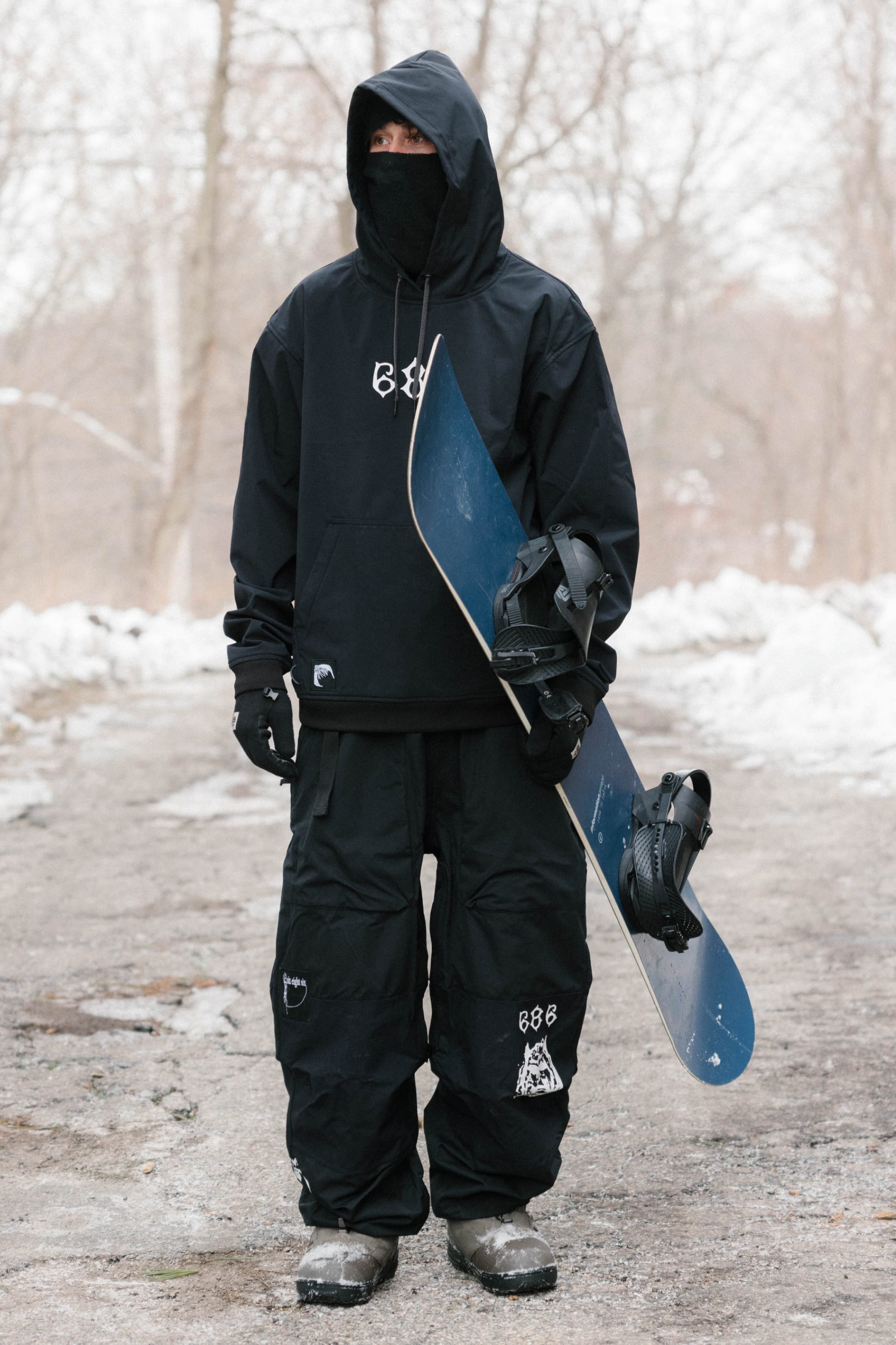A man stands in the snow wearing the 686 Mens Waterproof Team Hoody by 686, tan pants, and a black beanie with red and grey stripes. Snowy steps in the background create a mountain atmosphere.