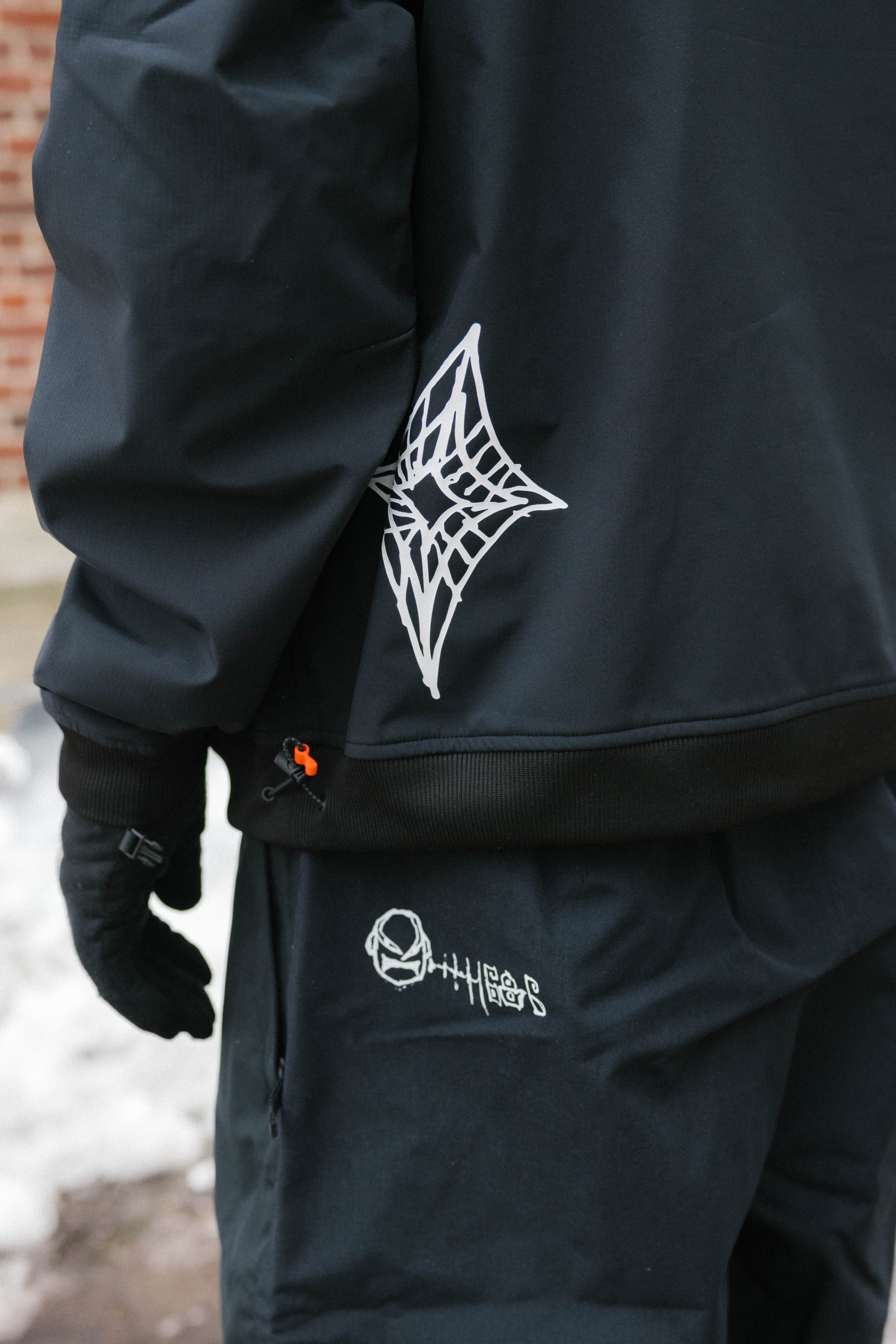 A person stands with their back to the camera against a white background, wearing a black beanie, 686 Mens Waterproof Team Hoody (black with 686 on the hood), and camouflage pants.