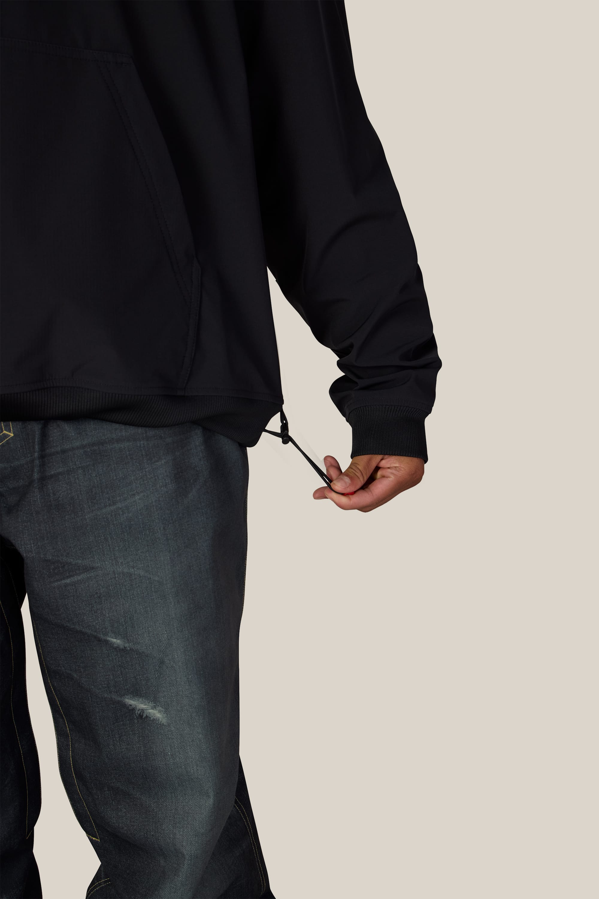 A person wearing the 686 Men’s Waterproof Team Hoody in black and distressed jeans adjusts the drawstring at the hem of the hoody. The background is plain beige.