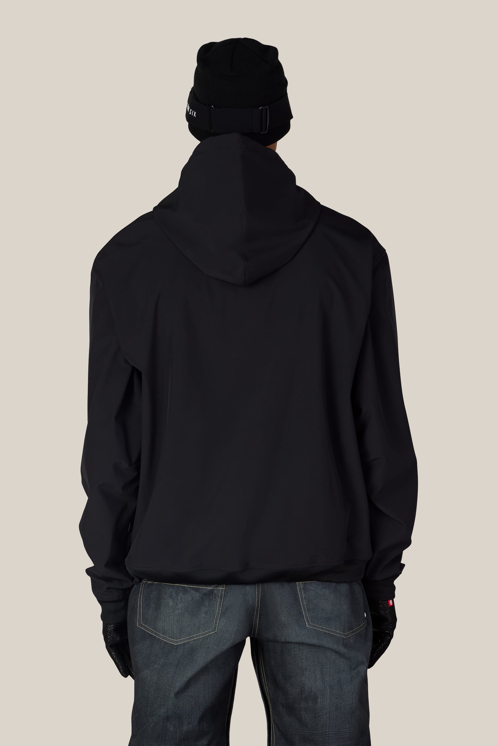 A person is standing with their back to the camera, wearing a 686 Men’s Waterproof Team Hoody in black, paired with blue jeans, black gloves, and a black beanie against a plain light background.
