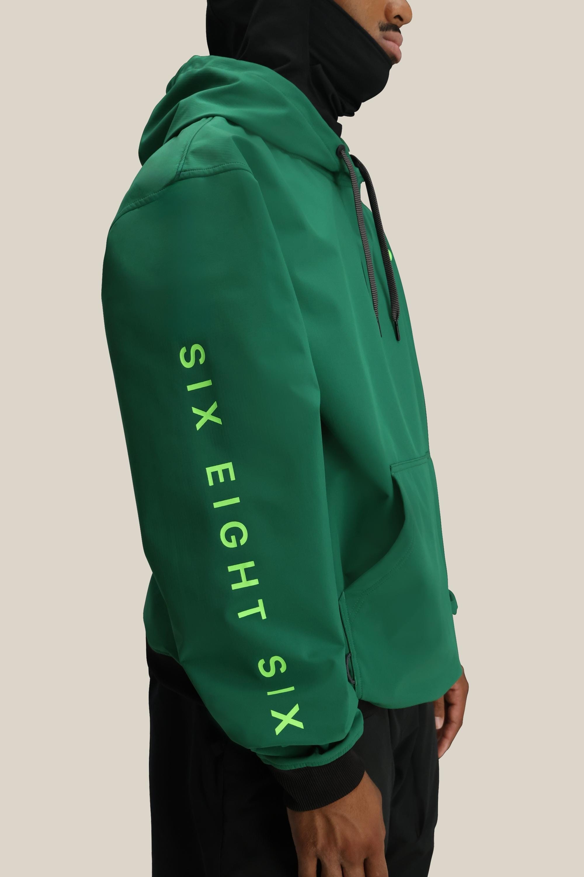 A person wearing a black 686 Mens Waterproof Team Hoody holds a yellow payphone receiver to their ear; only the lower face is visible, with a metal cord and “68a” sticker on the chest evoking a cool, mountain-inspired vibe.