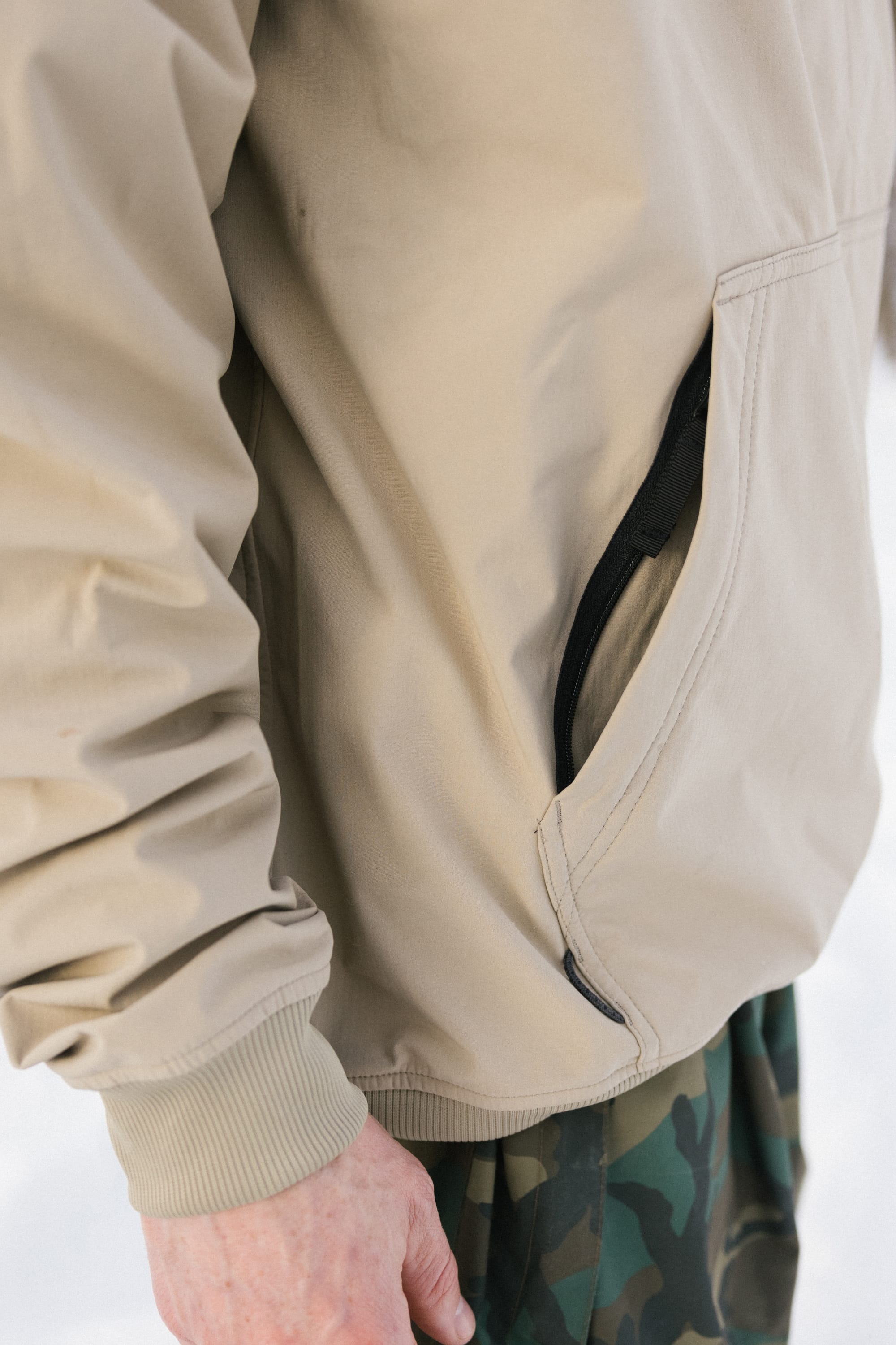 A person wearing the 686 Men’s Waterproof Team Hoody in beige, with their left hand near a zippered pocket and paired with camouflage pants; the background appears out of focus and light-colored.