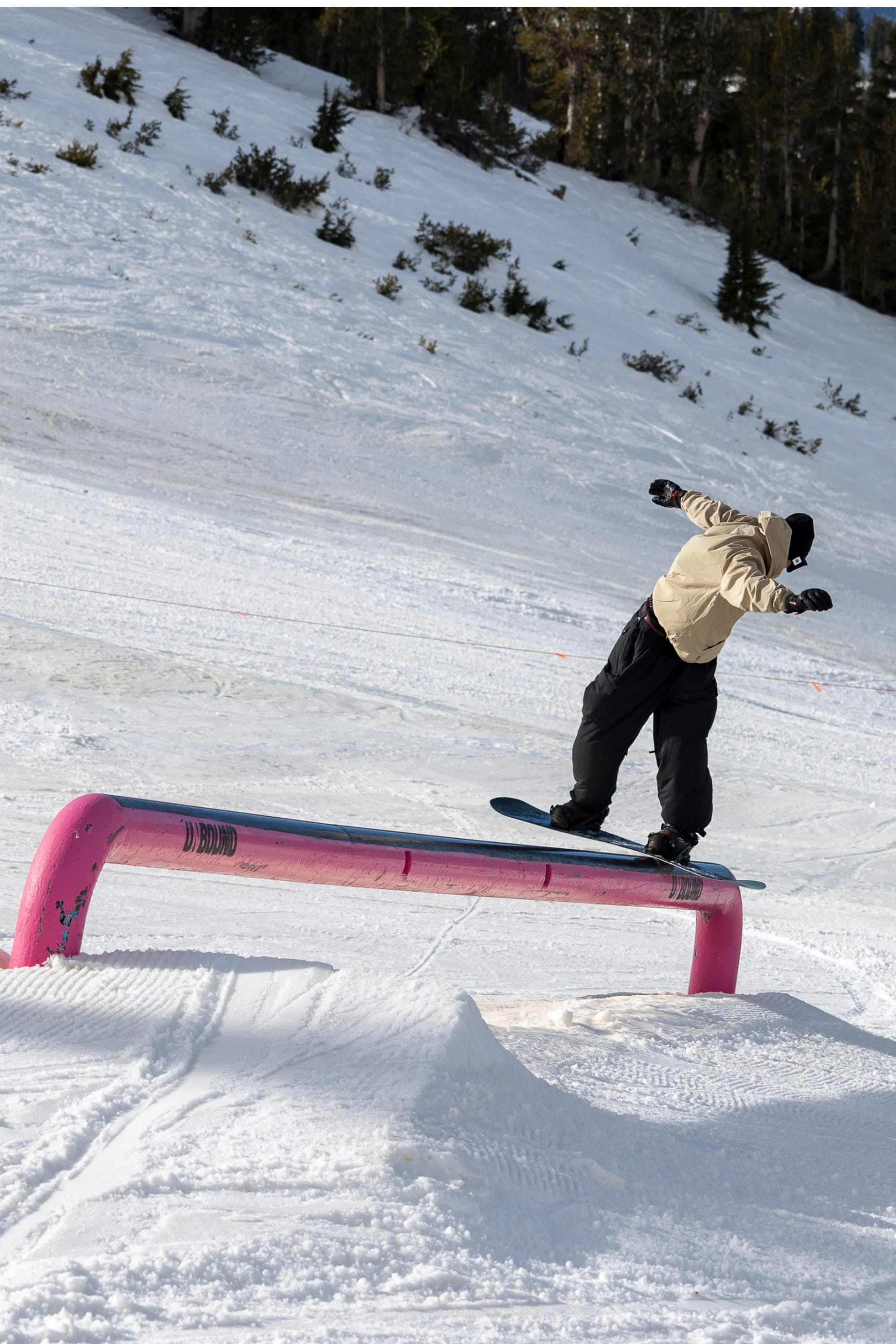 Wearing the 686 Men’s Waterproof Team Hoody, a snowboarder glides along a bright pink rail in a snowy terrain park, surrounded by snow and trees in the background.