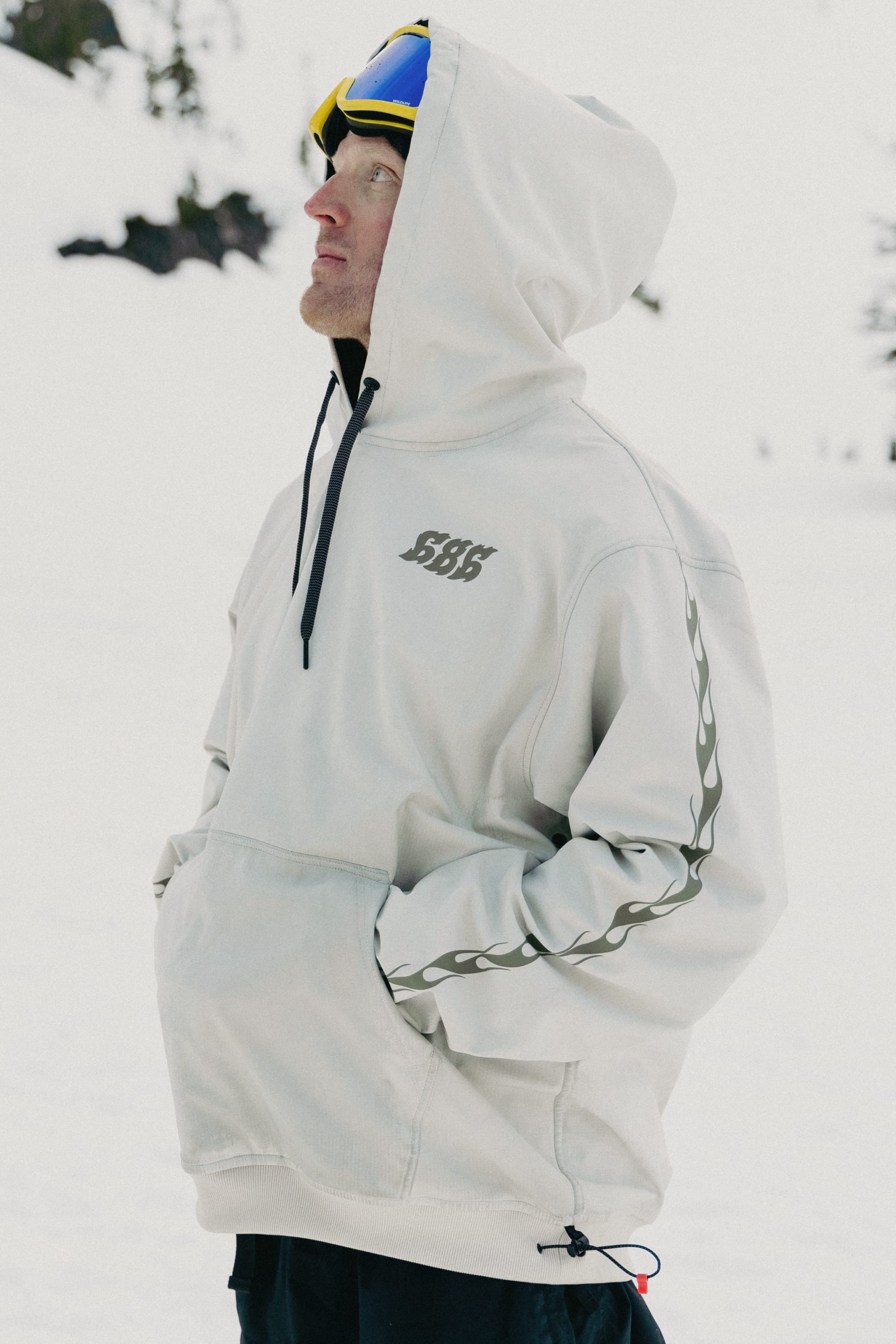 A person in a 686 Mens Waterproof Team Hoody with 888-HUF on the back stands outside in snowy mountains. The black-and-white, blurred photo highlights the advanced waterproof jacket by 686, built for harsh winter conditions.