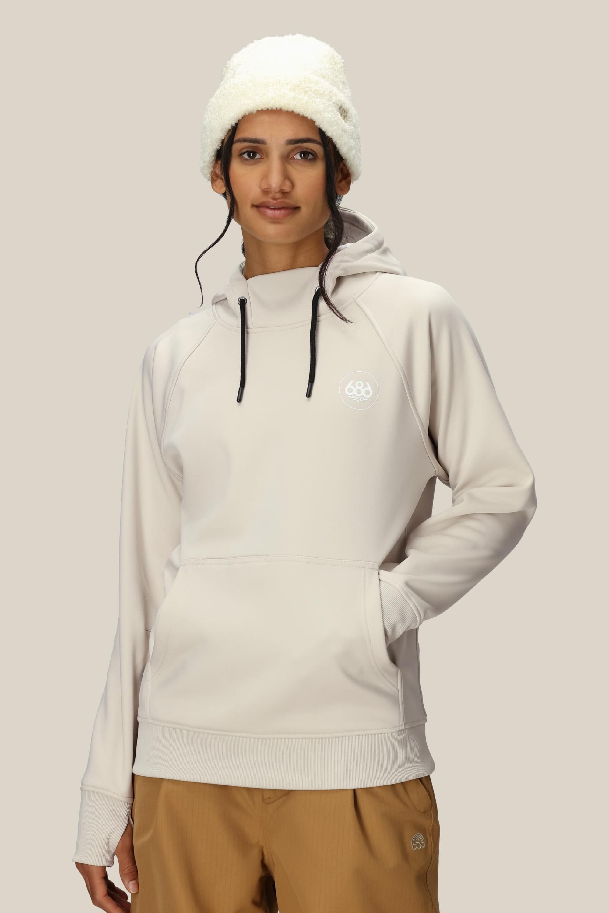 A woman in a cream-colored 686 Womens Bonded Fleece Pullover Hoody with a chest logo, tan pants, and a fluffy white hat stands against a beige background, looking at the camera with a neutral expression.
