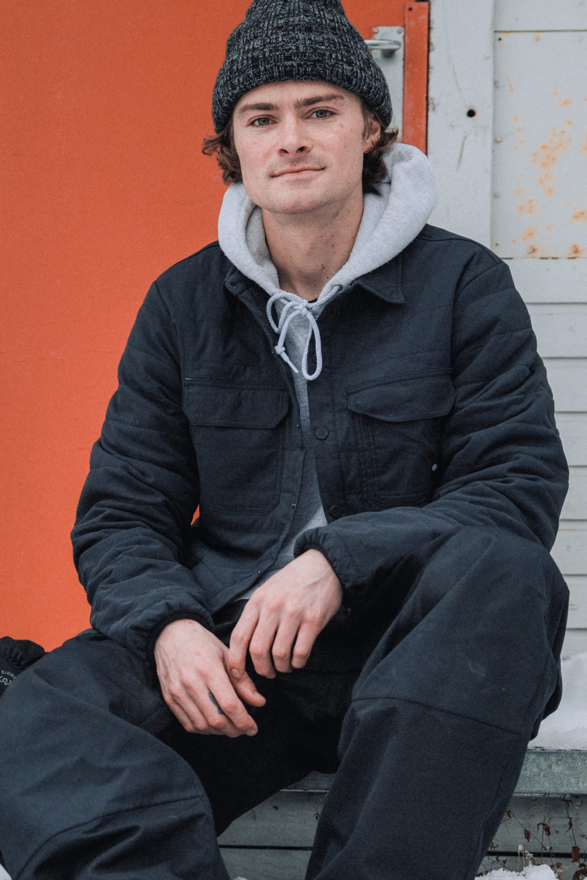 A young man in the 686 Mens Thermaldry Snap-Up Shirt sits outside against an orange and white wall, looking at the camera with a slight smile—effortlessly showcasing 686 style and performance in a modern city scene.