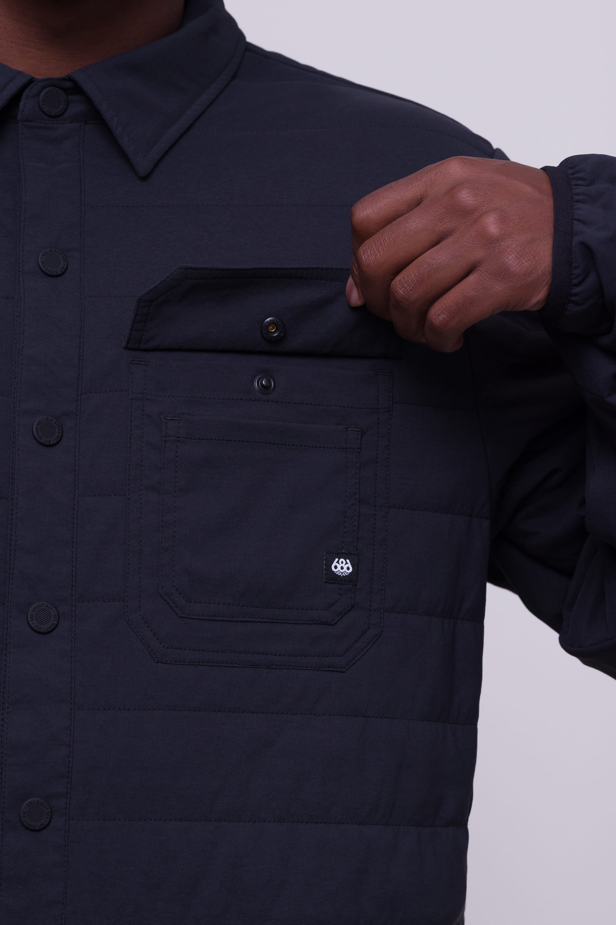 Wearing the 686 Mens Thermaldry Snap-Up Shirt, a person lifts the flap of a chest pocket to show an additional snap-button closure. Only their torso and one hand, clad in this performance shirt by 686, are visible.