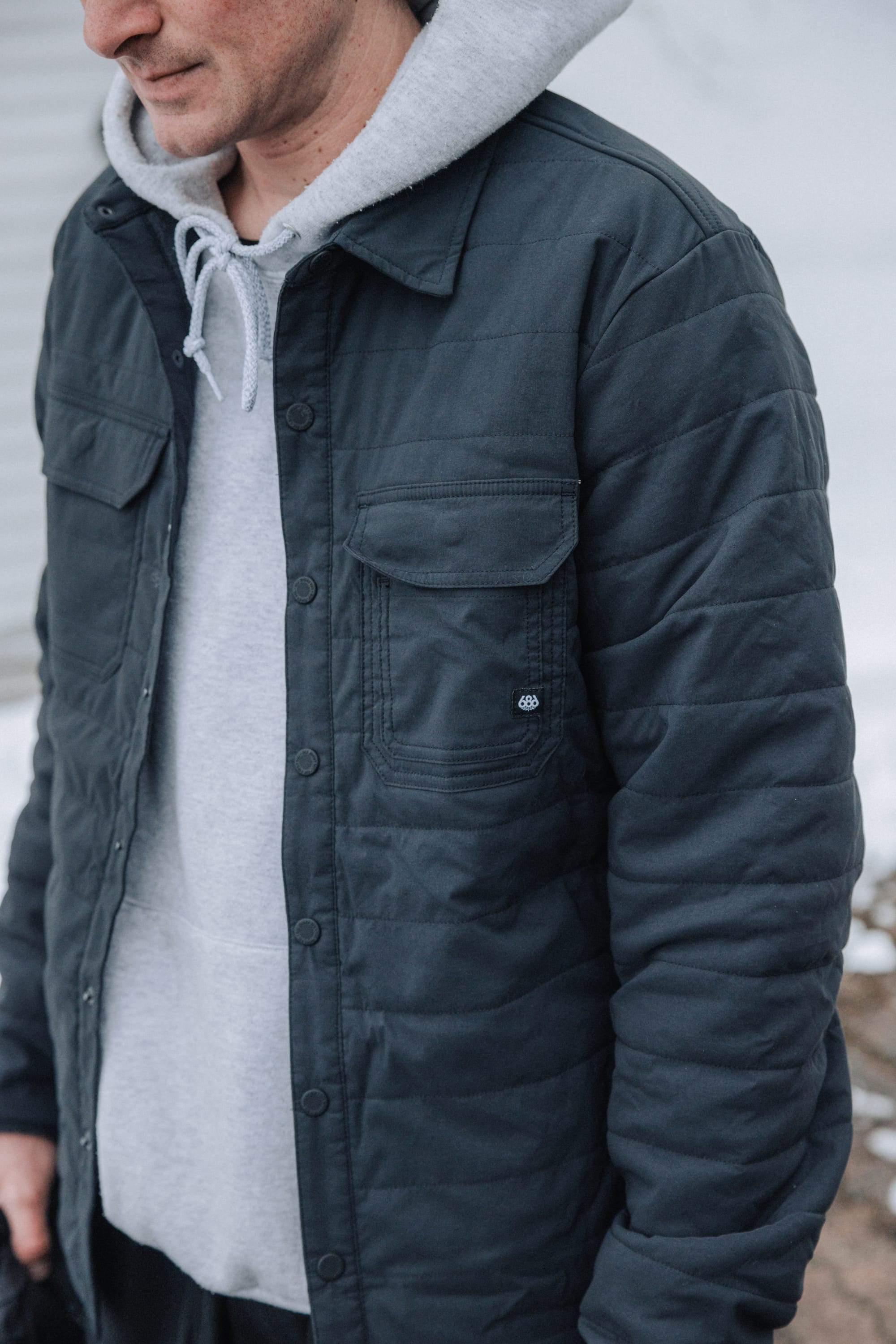 Wearing the 686 Mens Thermaldry Snap-Up Shirt, a person stands outdoors against a neutral city backdrop, partially visible from the shoulders down, channeling understated performance and urban style.