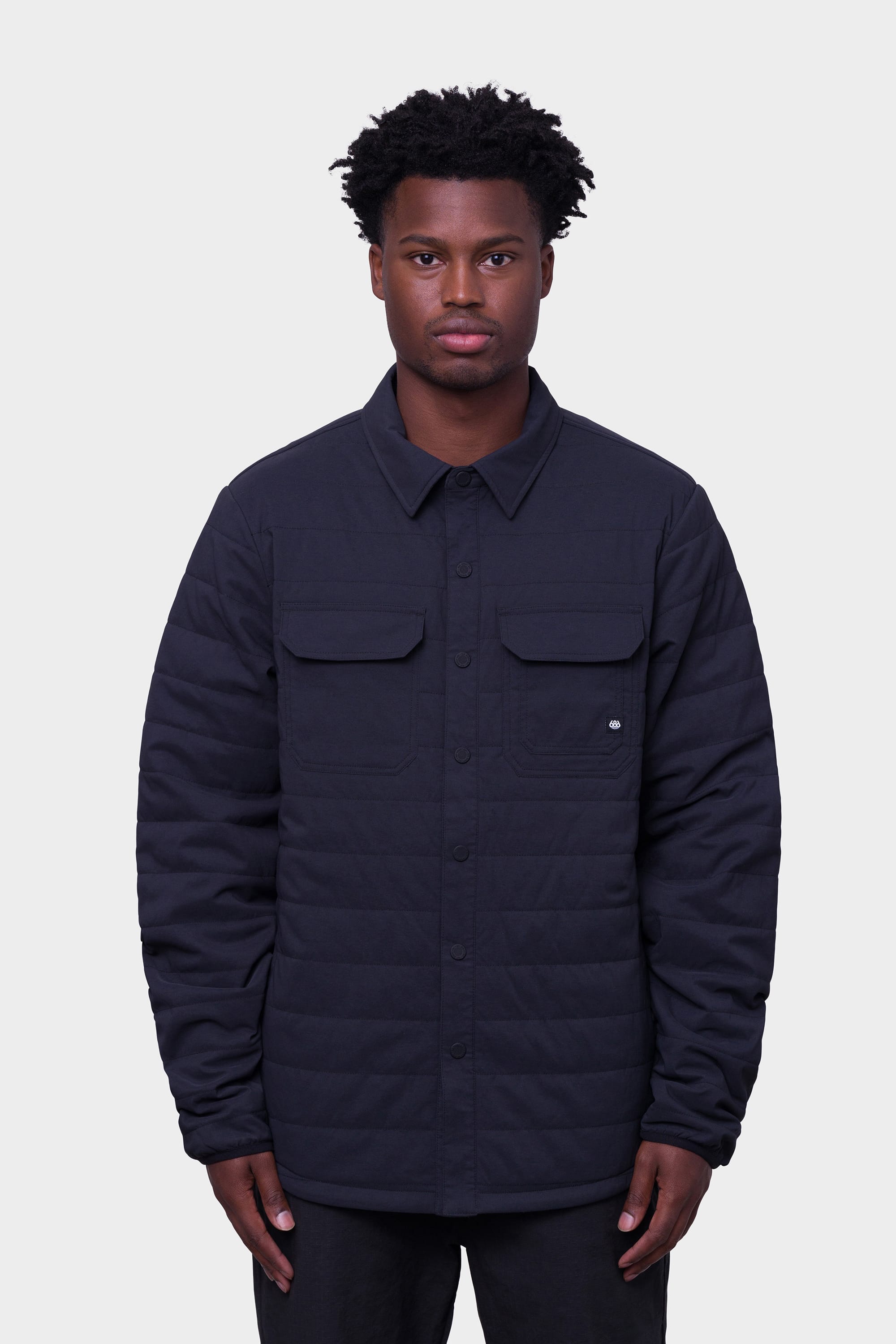 A man stands facing forward against a plain light gray background, wearing the 686 Mens Thermaldry Snap-Up Shirt—a black merino-blend quilted button-up with two chest pockets from 686.