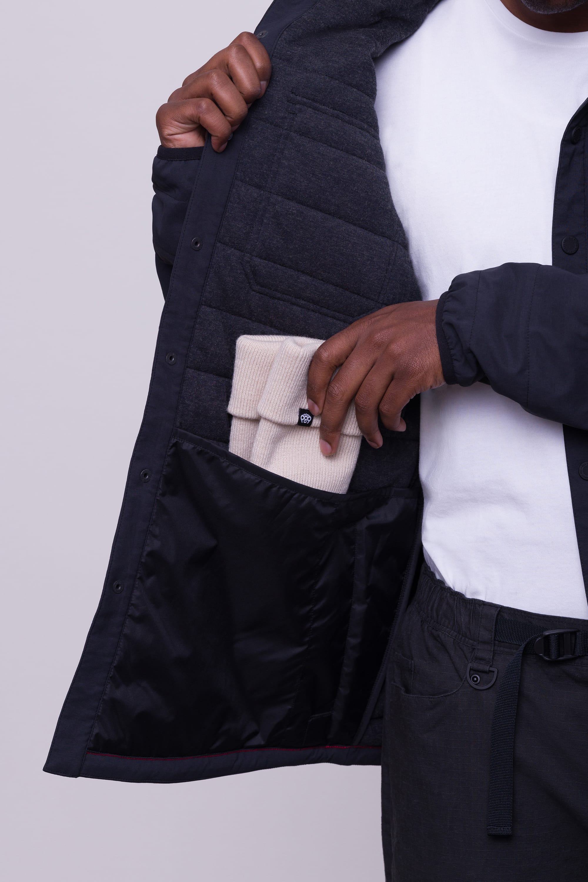 A man wearing the 686 Mens Thermaldry Snap-Up Shirt opens his jacket to reveal an inner pocket, placing a folded beige merino blend knit hat inside—showcasing 686’s understated performance for stylish city dwellers.