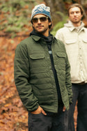 Two men stand in a forest among fallen leaves; the man in front wears the 686 Mens Thermaldry Snap-Up Shirt with sunglasses and a beanie, embodying understated performance, while the man behind him sports a light-colored quilted jacket.