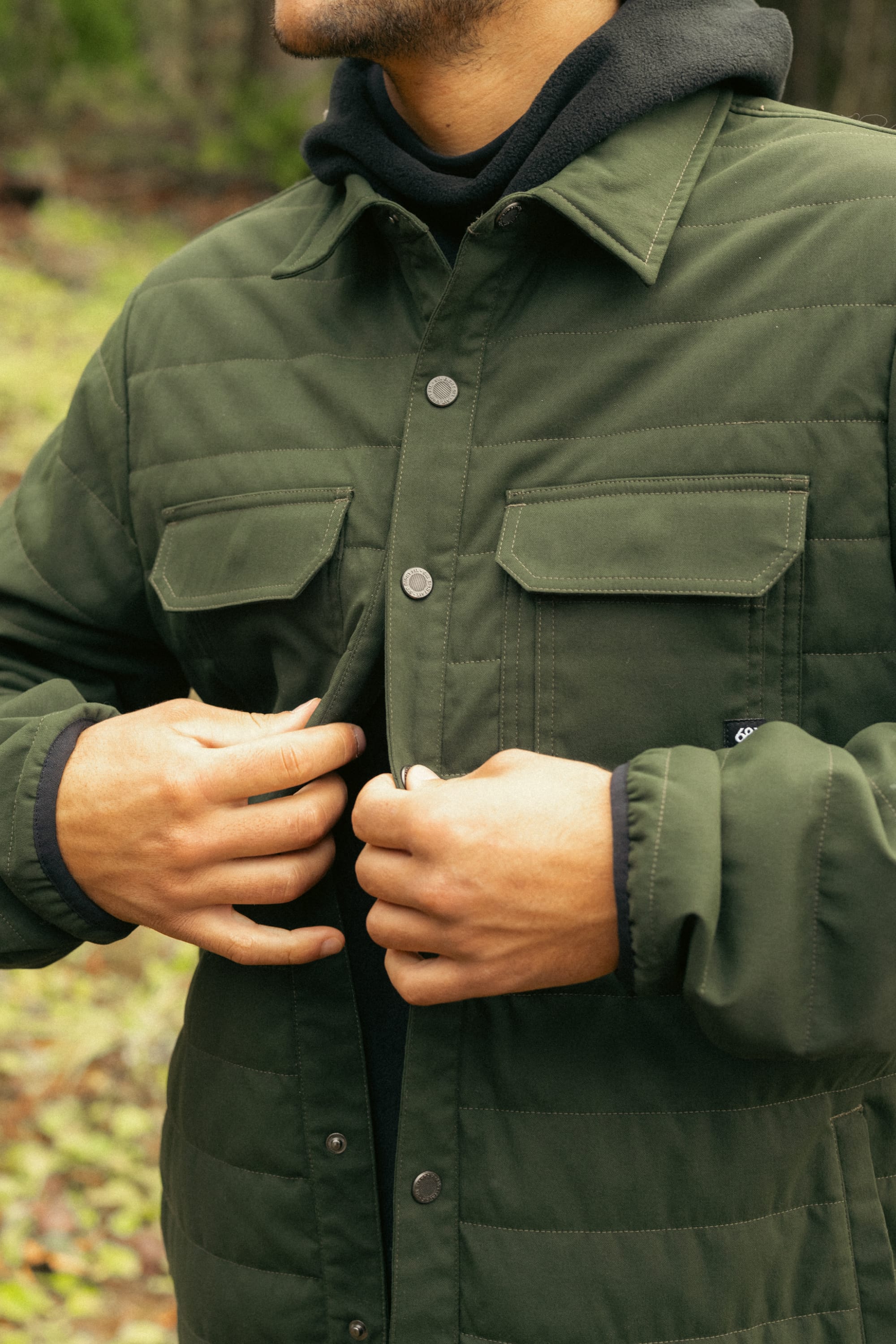 Wearing the 686 Mens Thermaldry Snap-Up Shirt in green over a black hoodie, a person fastens the snaps—showcasing 686’s modern performance style against an urban backdrop. The outdoor photo has their face partially out of frame.