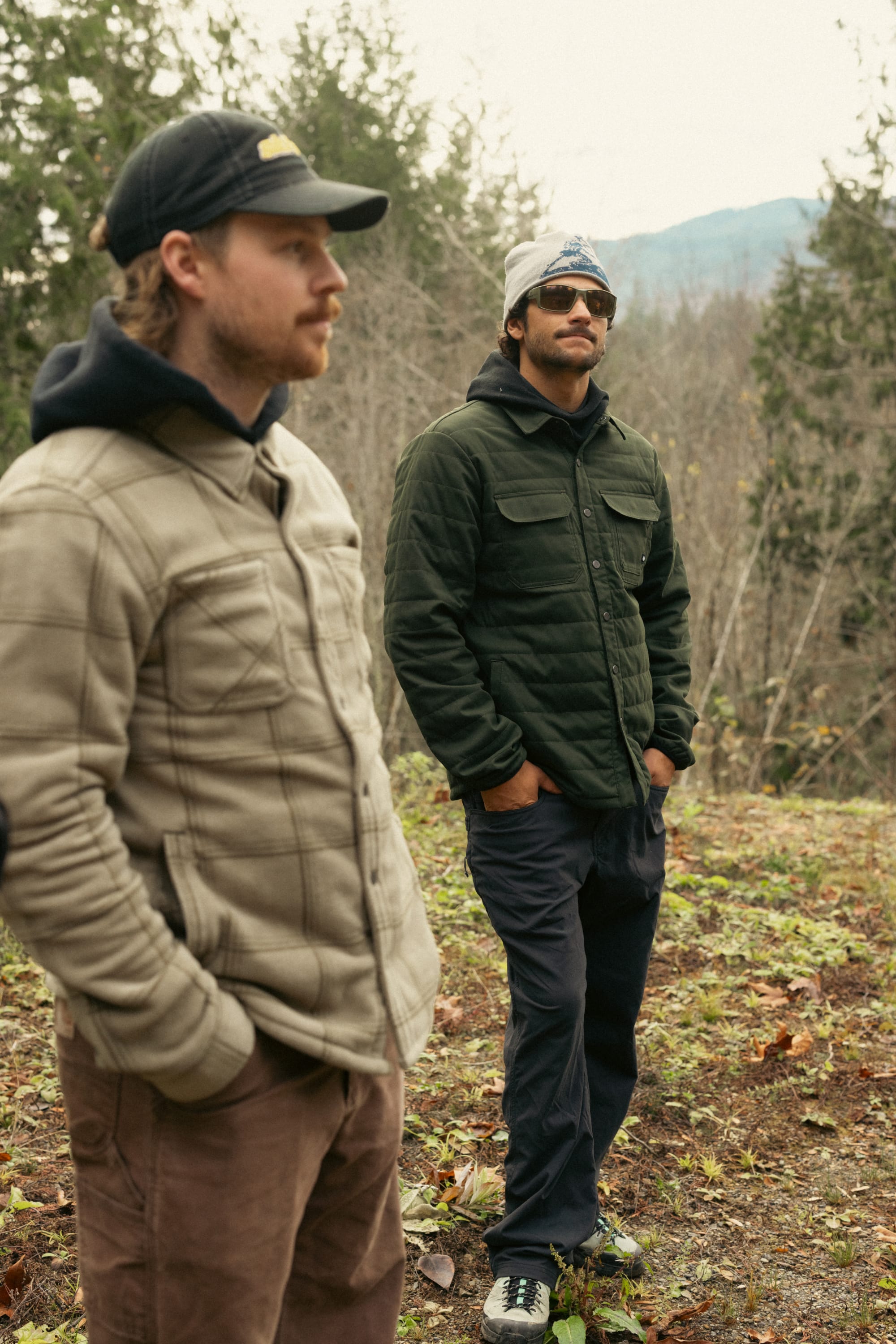 Two men stand on grass outdoors; one wears a tan jacket and black cap, the other wears sunglasses, a bandana, and a dark green 686 Mens Thermaldry Snap-Up Shirt. Trees and distant mountains complete the subtle performance scene.