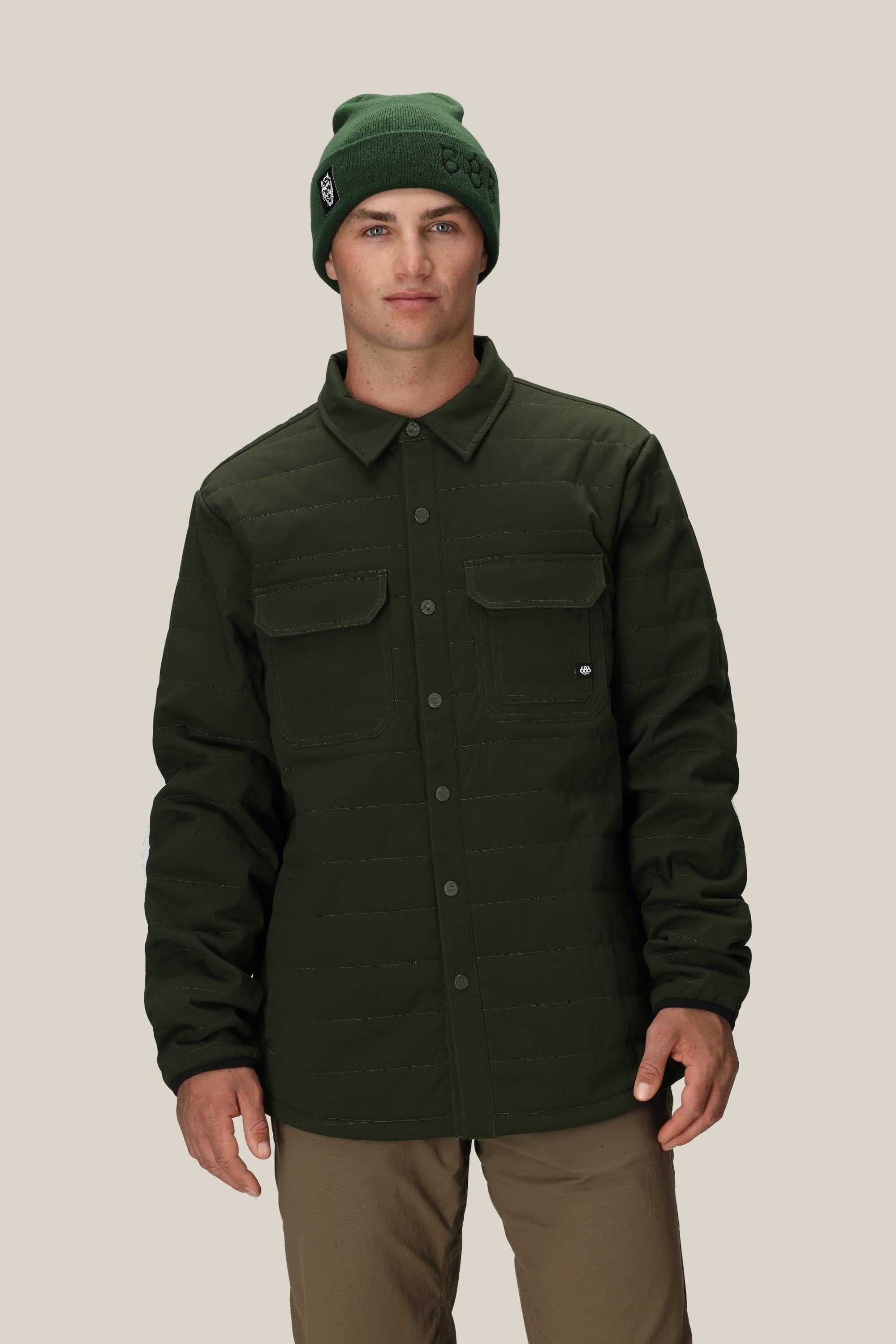 A man stands facing forward against a beige background, wearing the 686 Mens Thermaldry Snap-Up Shirt in dark green, a matching beanie, and tan pants—delivering understated performance and style by 686 for urban settings.