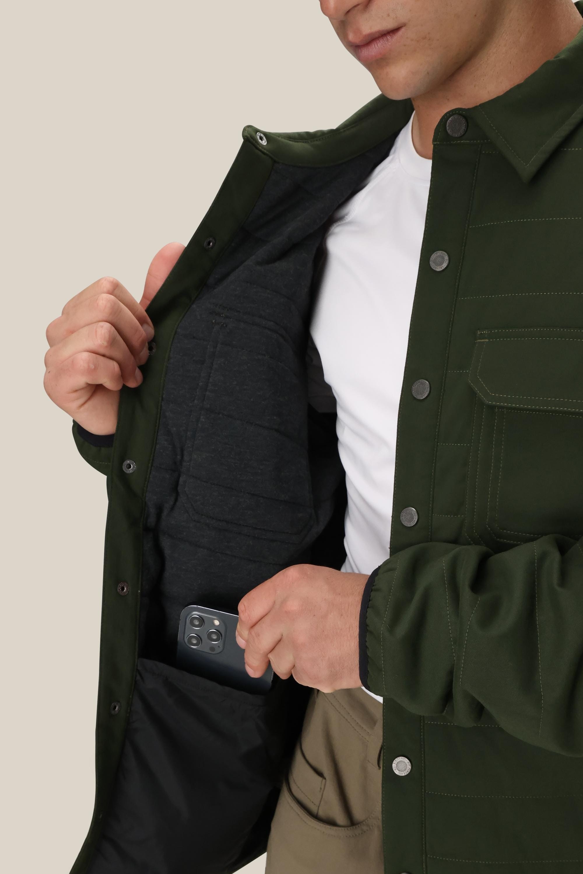 A man in the 686 Mens Thermaldry Snap-Up Shirt, paired with tan pants, opens his green jacket to reveal a spacious inner pocket and slips in his smartphone—showcasing discreet utility and urban style from 686.