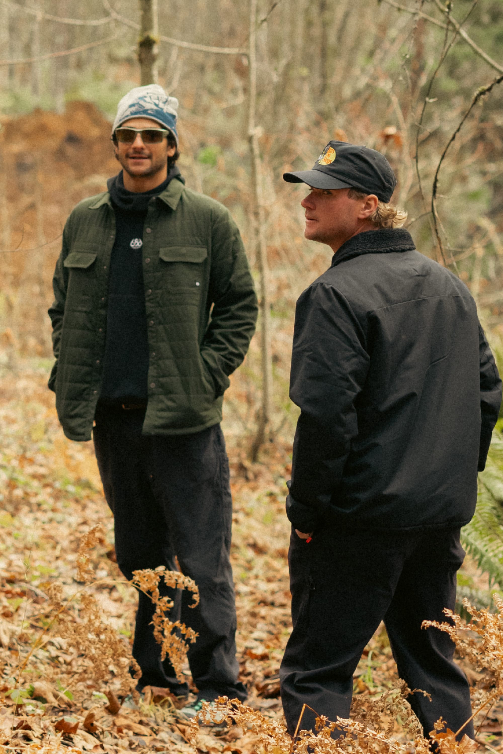Two men stand in an autumn forest wearing 686 Mens Thermaldry Snap-Up Shirts. One pairs his green shirt with a blue hat; the other opts for black with a cap. Both appear relaxed among the trees and fallen leaves.