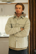 A man with light brown hair and a short beard stands indoors with arms crossed, modeling the 686 Mens Thermaldry Snap-Up Shirt over a hoodie—an image of understated performance in a stylish city setting.