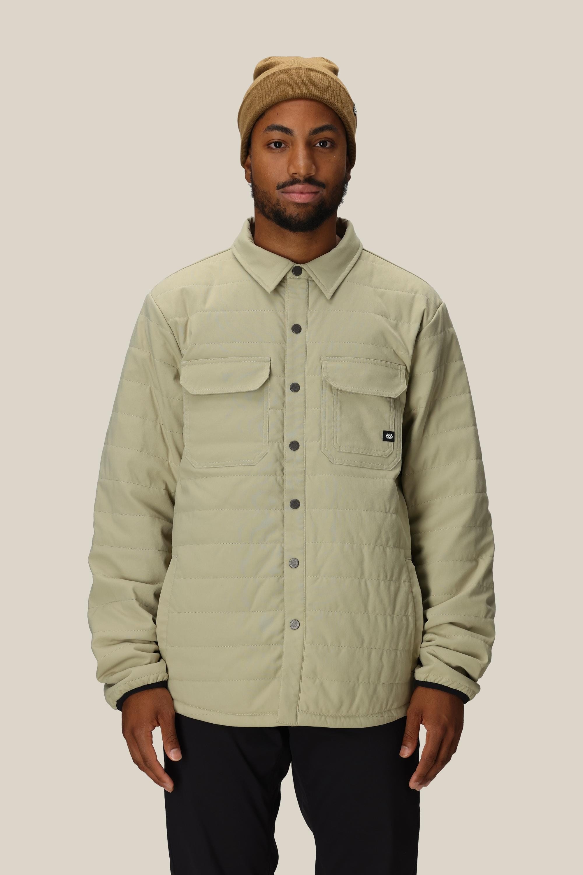 A man in the 686 Mens Thermaldry Snap-Up Shirt—tan, quilted, with snap buttons and two chest pockets—paired with black pants and a tan beanie stands against a light beige background, reflecting understated urban style.