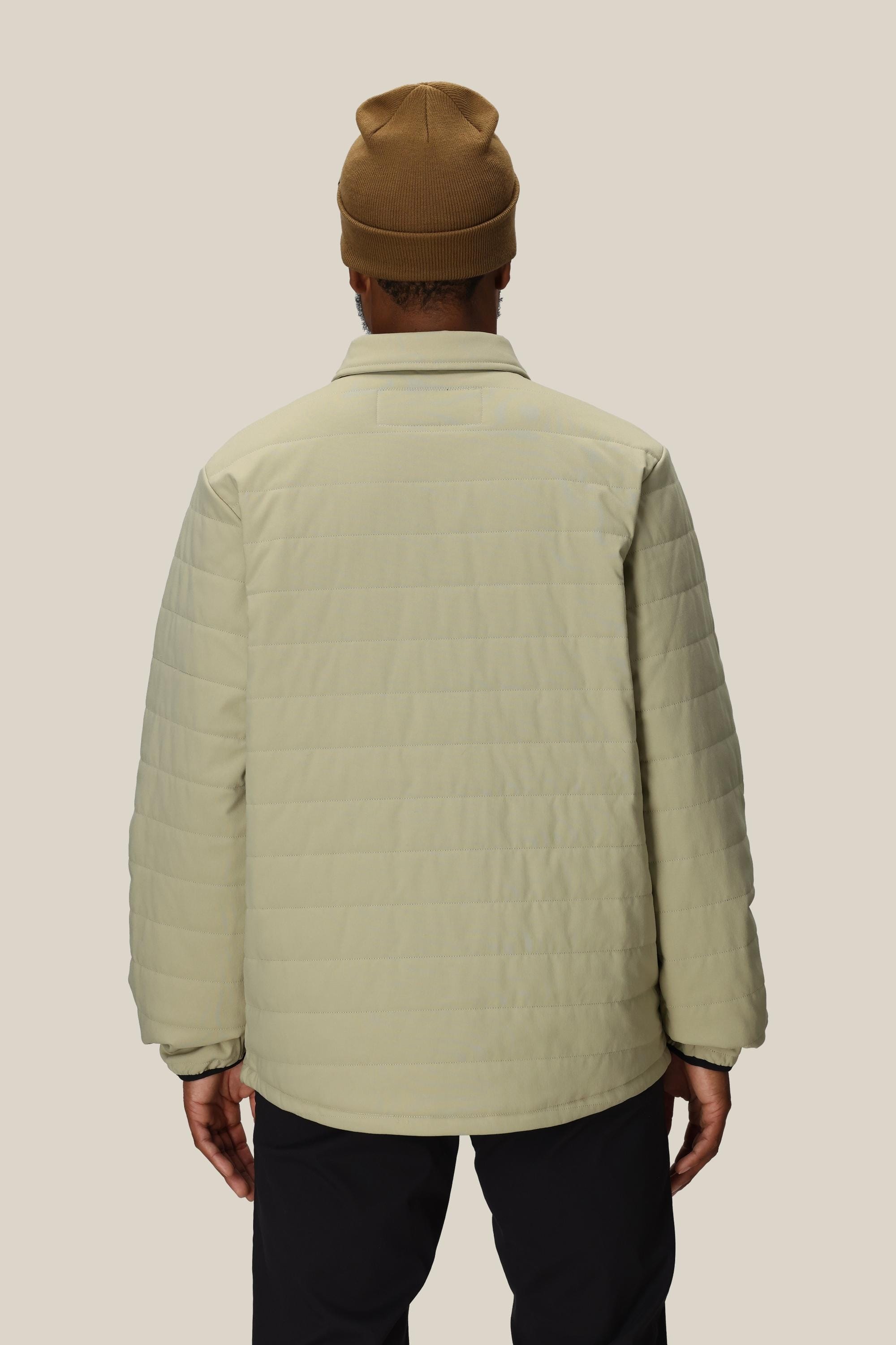 A person stands facing away, wearing the 686 Mens Thermaldry Snap-Up Shirt in light beige, a brown knit beanie, and dark pants—a sleek urban look by 686 against a plain light background.