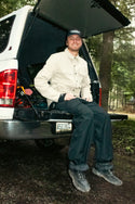 A smiling man wearing the 686 Mens Thermaldry Snap-Up Shirt sits on the open tailgate of a white van among forest trees, with his adventure-ready gear visible in the trunk, ready for understated outdoor performance.