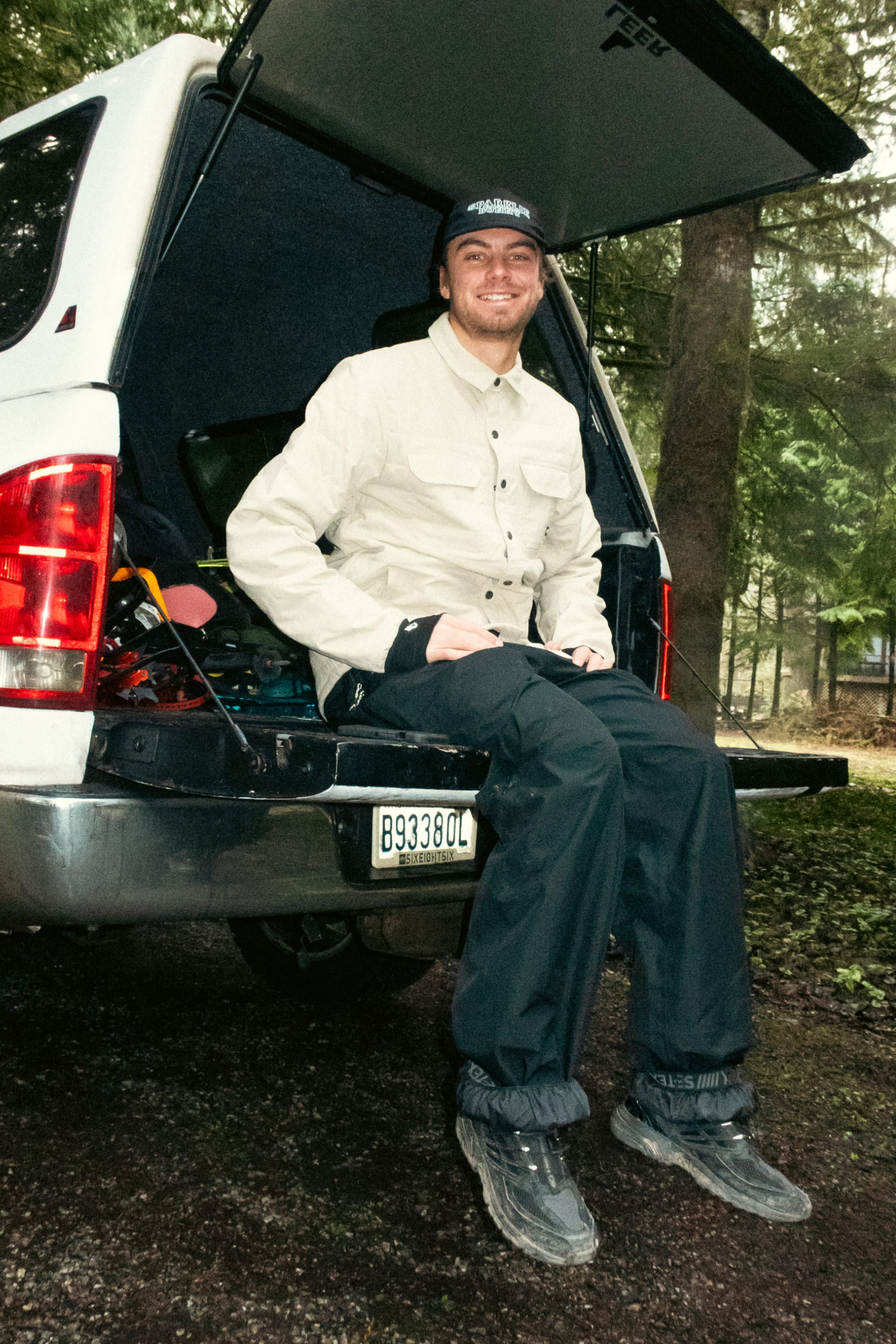 A smiling man wearing the 686 Mens Thermaldry Snap-Up Shirt sits on the open tailgate of a white van among forest trees, with his adventure-ready gear visible in the trunk, ready for understated outdoor performance.