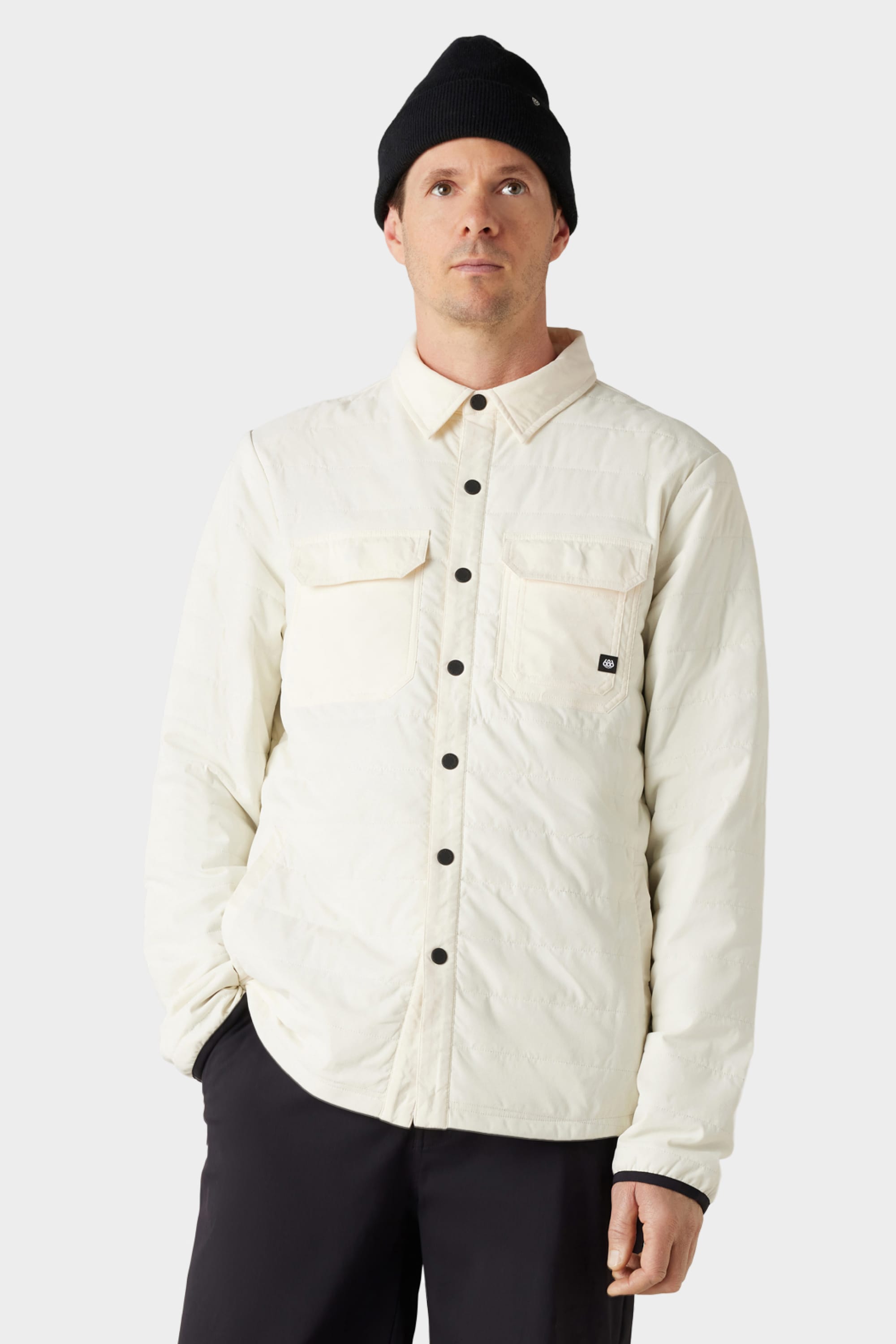 A man wears the 686 Mens Thermaldry Snap-Up Shirt, a white quilted merino blend with chest pockets by 686, styled with black pants and a black beanie against a plain light gray background for subtle city-ready performance.