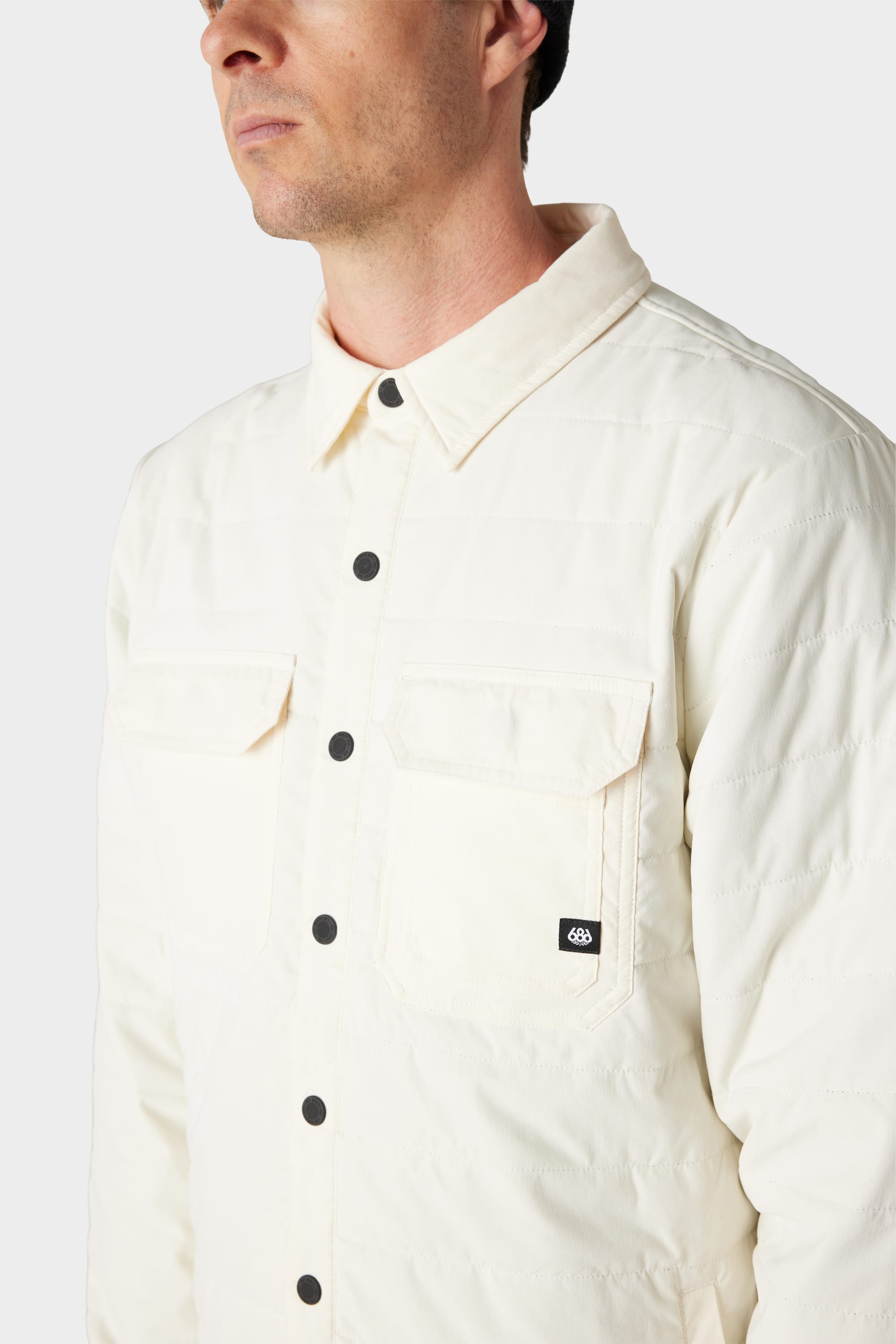 A man wears the 686 Mens Thermaldry Snap-Up Shirt—a cream merino blend quilted jacket with two chest pockets and black buttons—shown from the shoulders up against a plain background, ideal for understated city style.