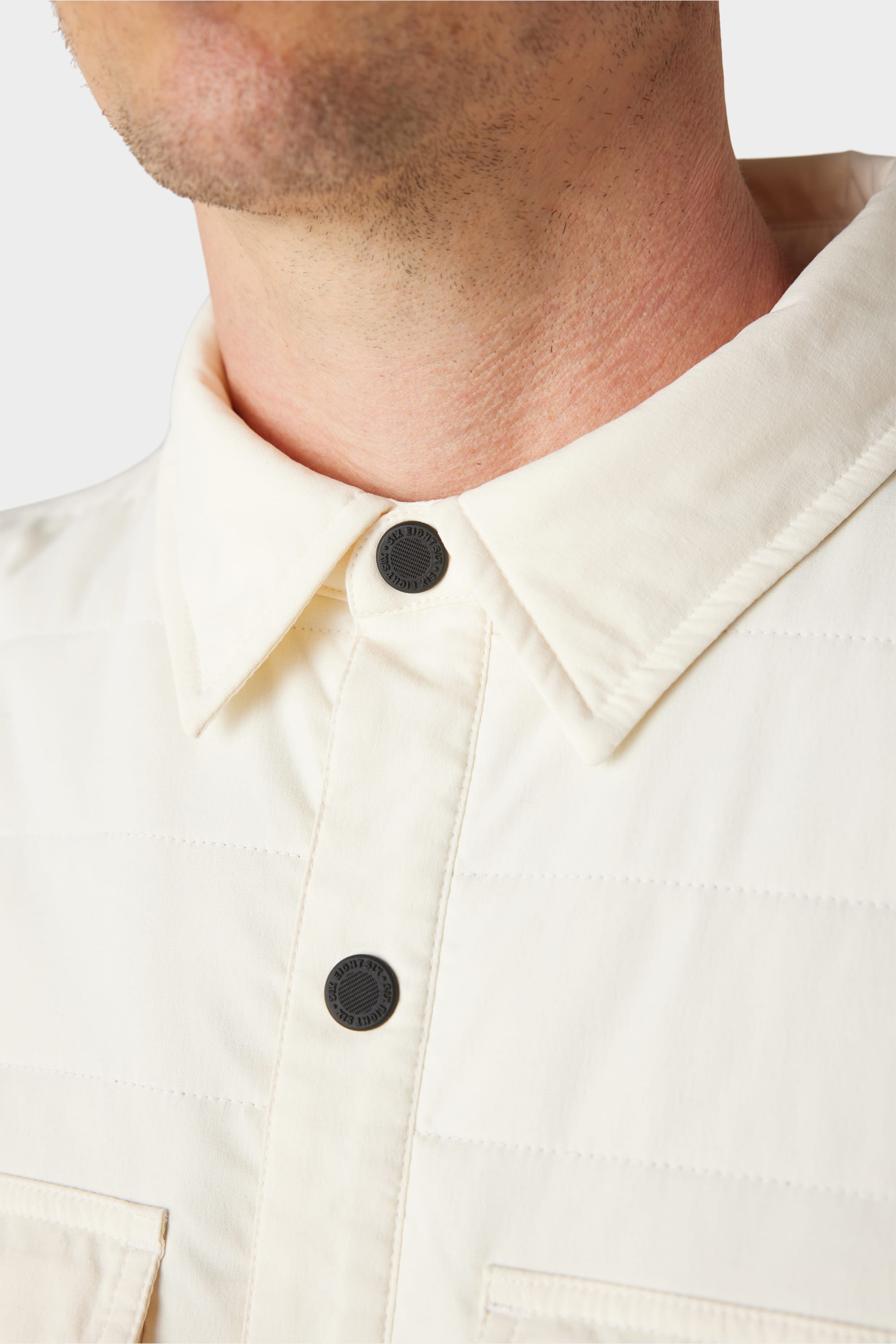 Close-up of a person wearing the 686 Men’s Thermaldry Snap-Up Shirt in cream, featuring black buttons and a collared design—an understated, stylish city look from 686. Only the lower half of their face and neck are visible.