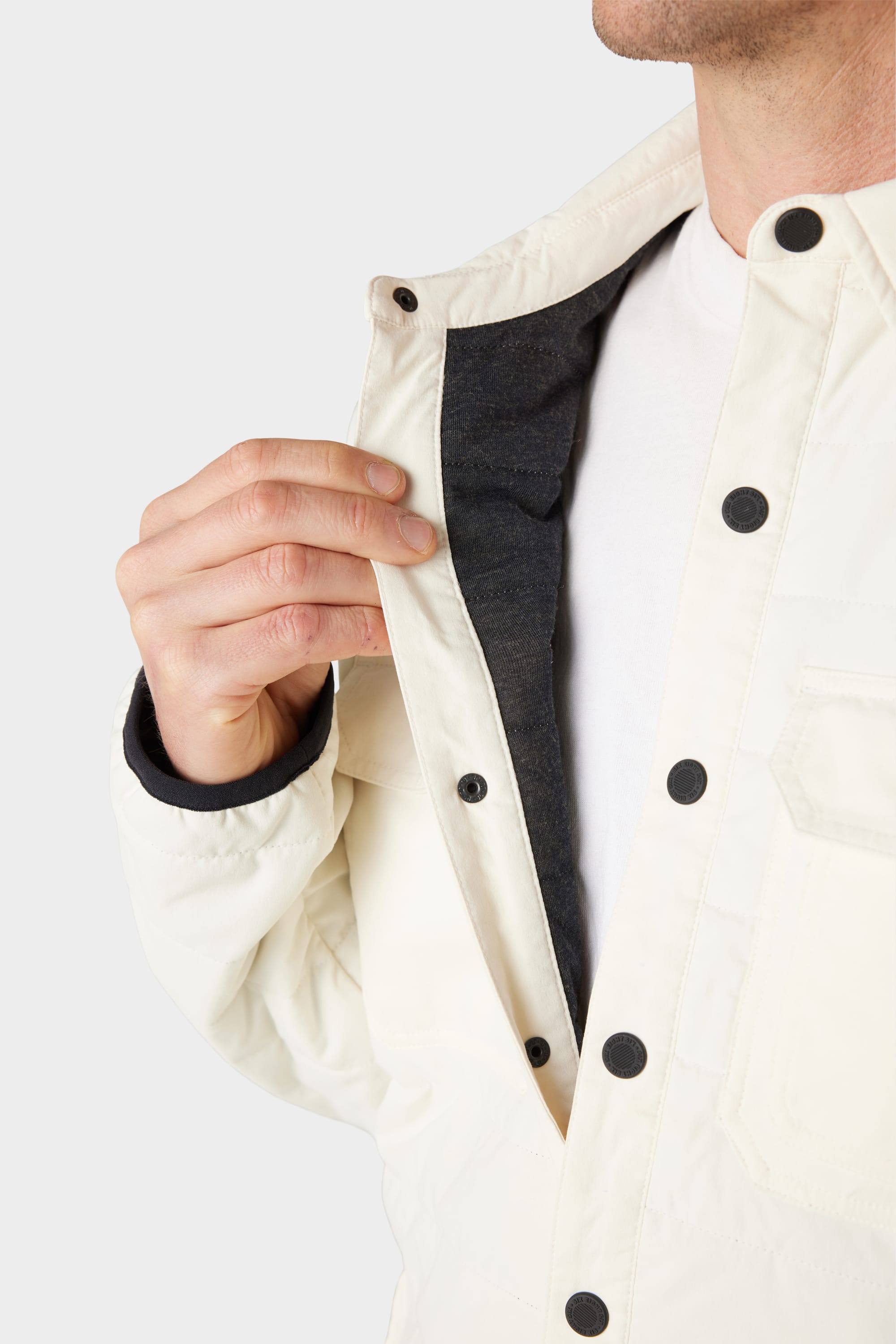 A person wearing the 686 Mens Thermaldry Snap-Up Shirt in white over a white shirt pulls open the jacket to reveal its dark inner lining. Only their lower face and upper body are visible.
