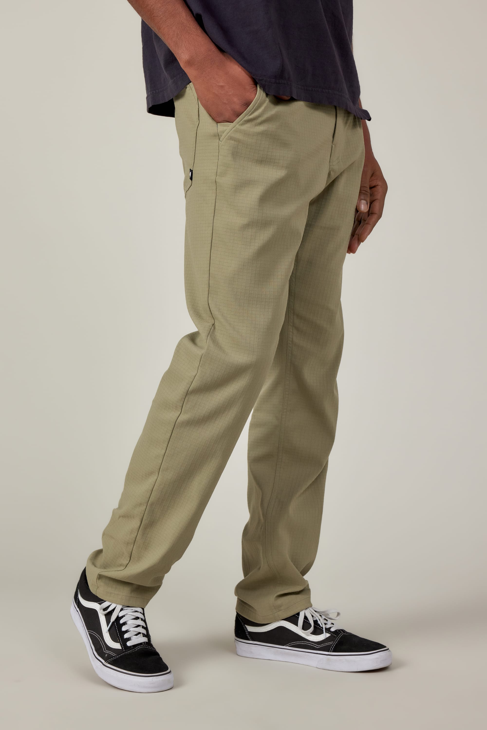 A person wearing 686 Mens Everywhere Ripstop Pant - Slim Fit in light olive, a dark t-shirt, and black Vans sneakers stands with one hand in their pocket against a plain background.