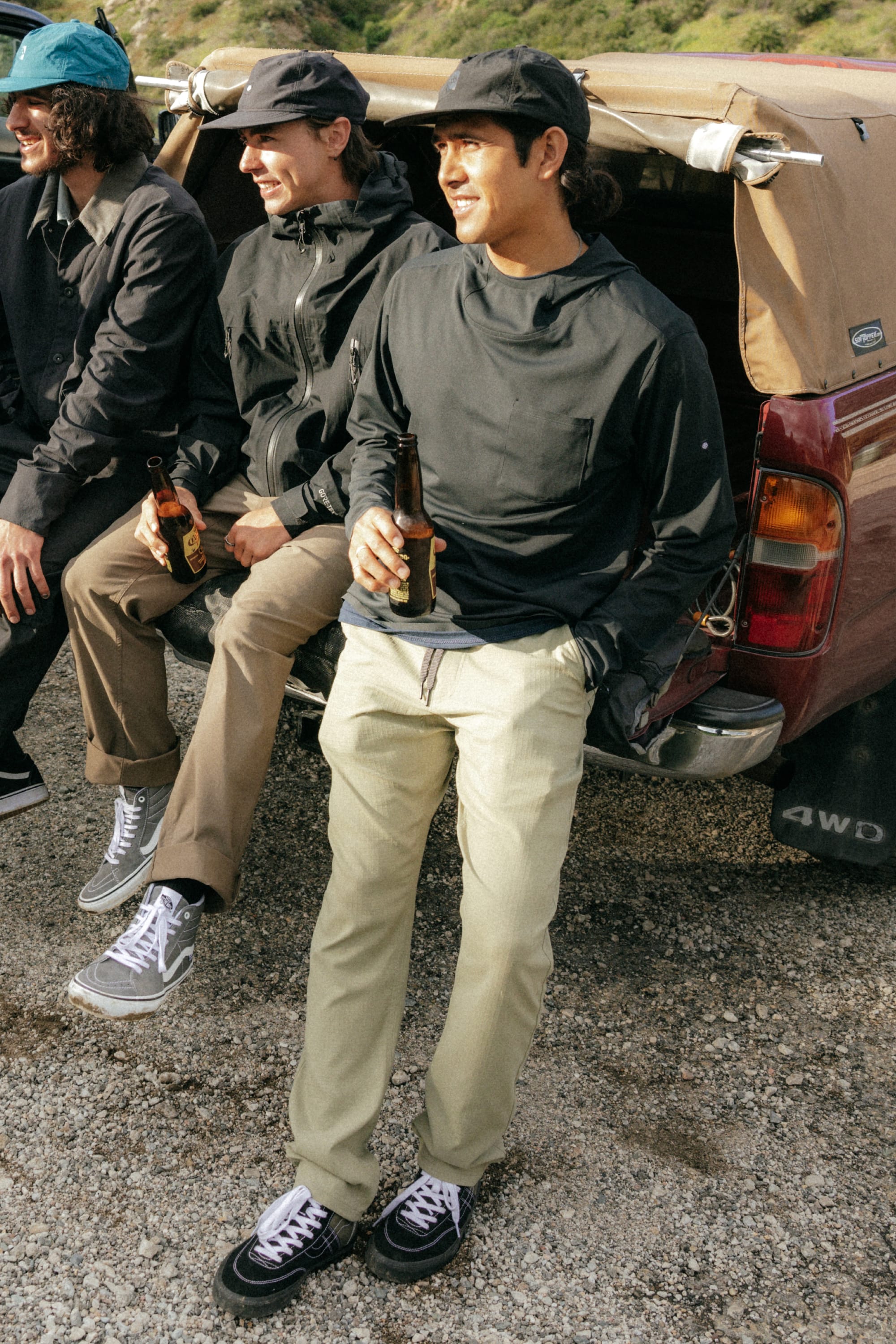 Three people in casual outdoor clothing, including 686 Mens Everywhere Ripstop Pant - Slim Fit by 686, lean against a vehicle holding beer bottles and smiling in a relaxed outdoor setting with sunlight and greenery.