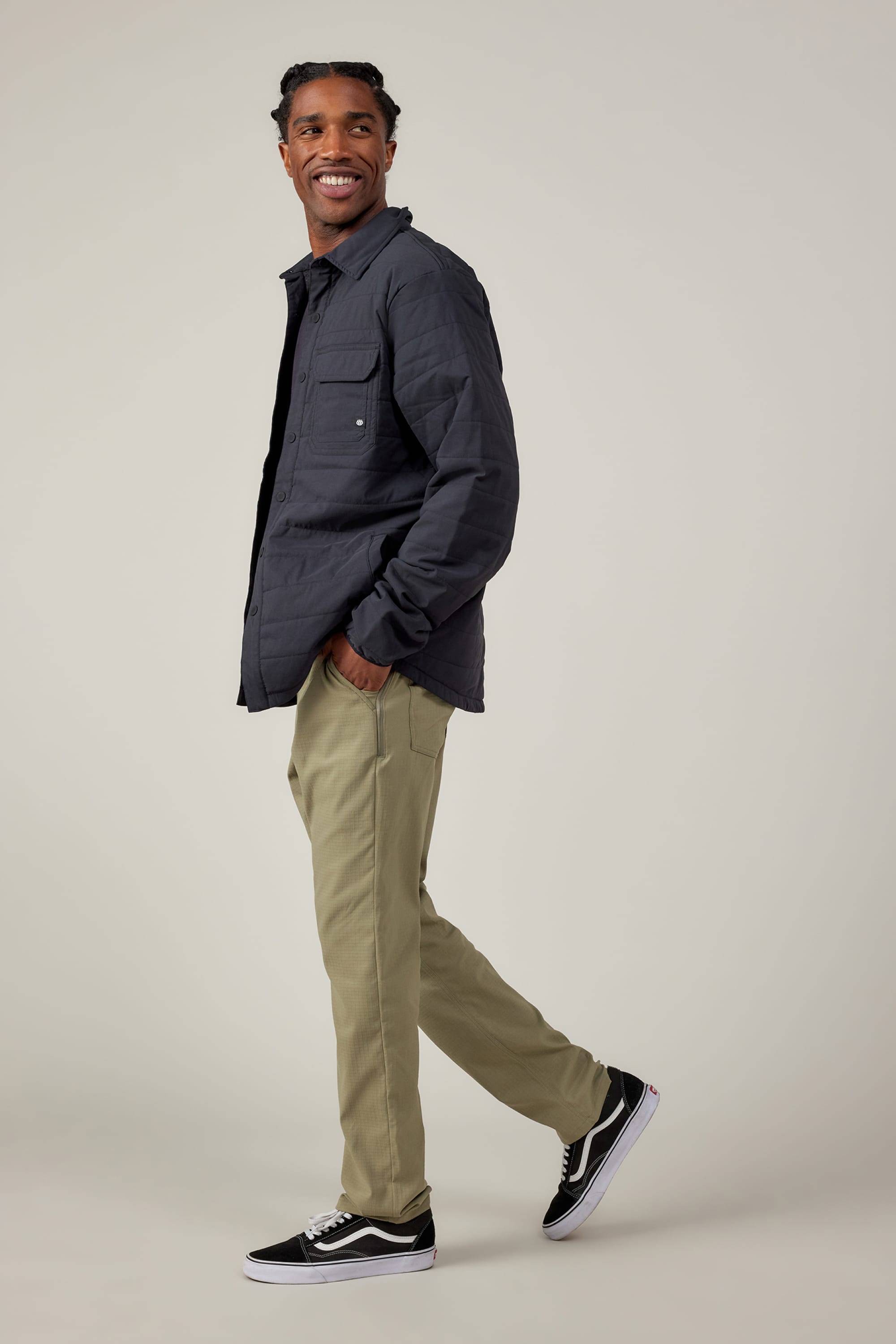 A man with braided hair smiles while standing sideways in 686 Mens Everywhere Ripstop Pant - Slim Fit by 686, paired with a dark quilted jacket and black-and-white sneakers against a plain light background.