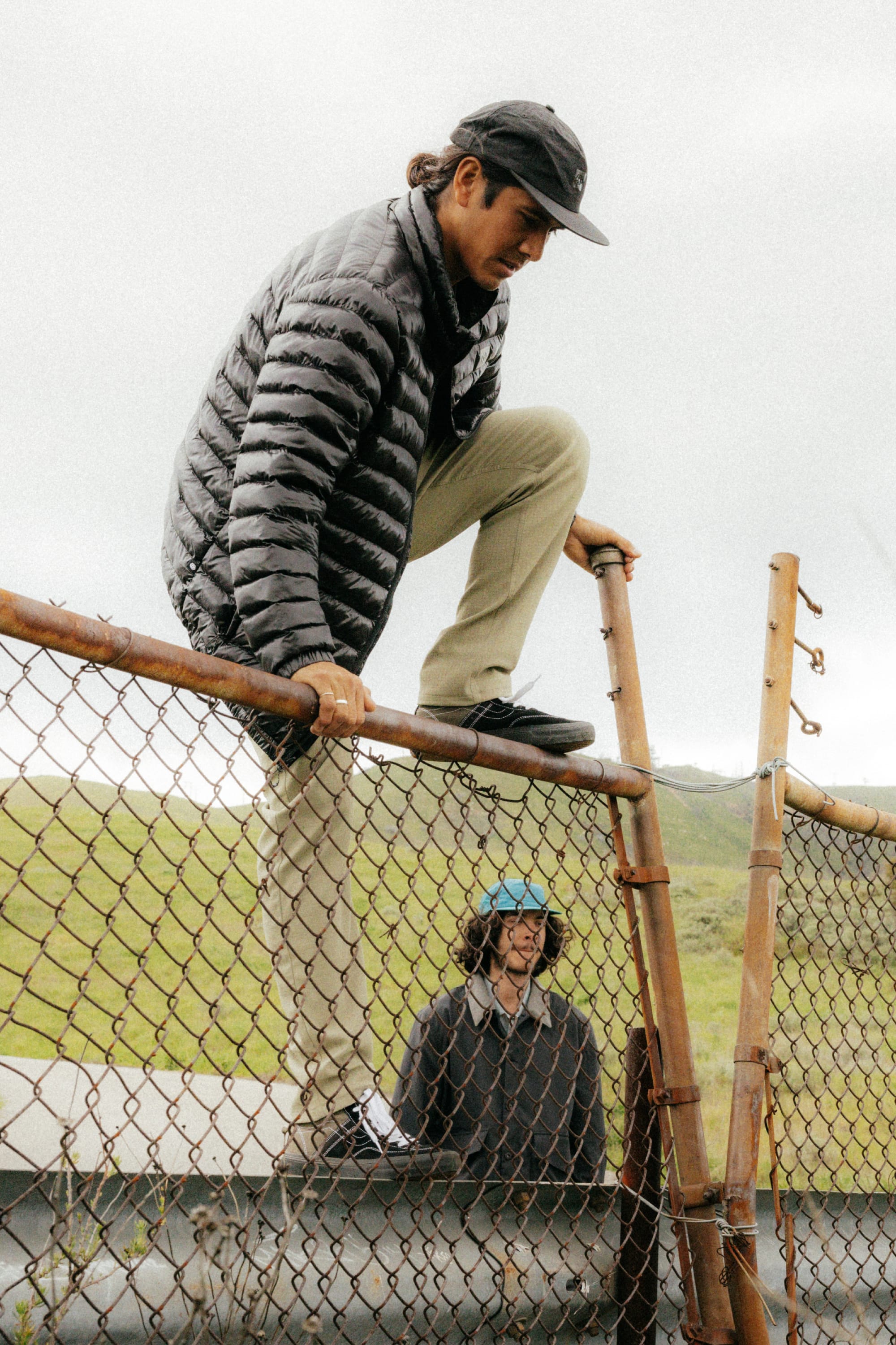 A man wearing 686 Mens Everywhere Ripstop Pant - Slim Fit climbs over a rusty chain-link fence, while another person in a helmet stands on the other side in a grassy outdoor area.