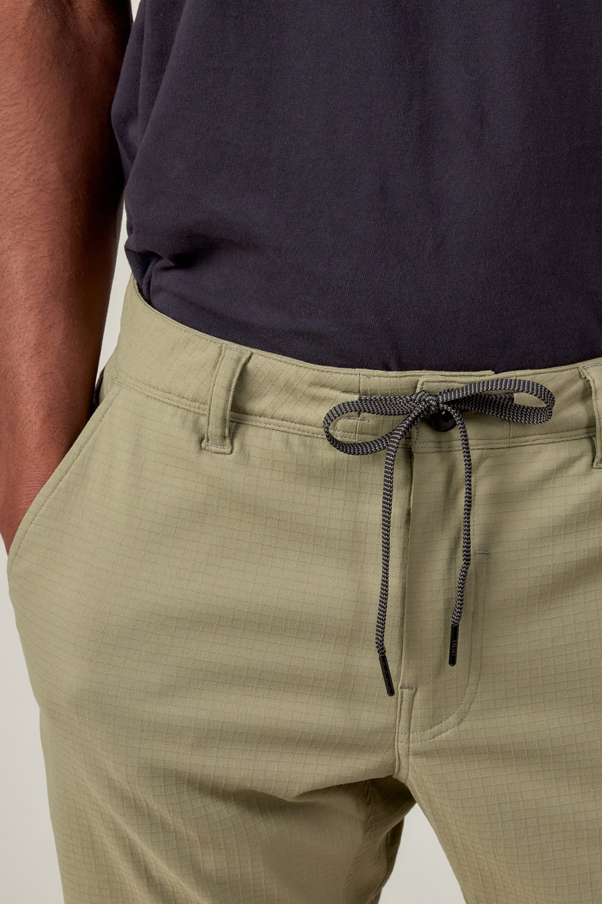 A person wears 686 Mens Everywhere Ripstop Pant - Slim Fit in light olive green with a black-and-white drawstring, paired with a dark t-shirt and their left hand in their pocket.