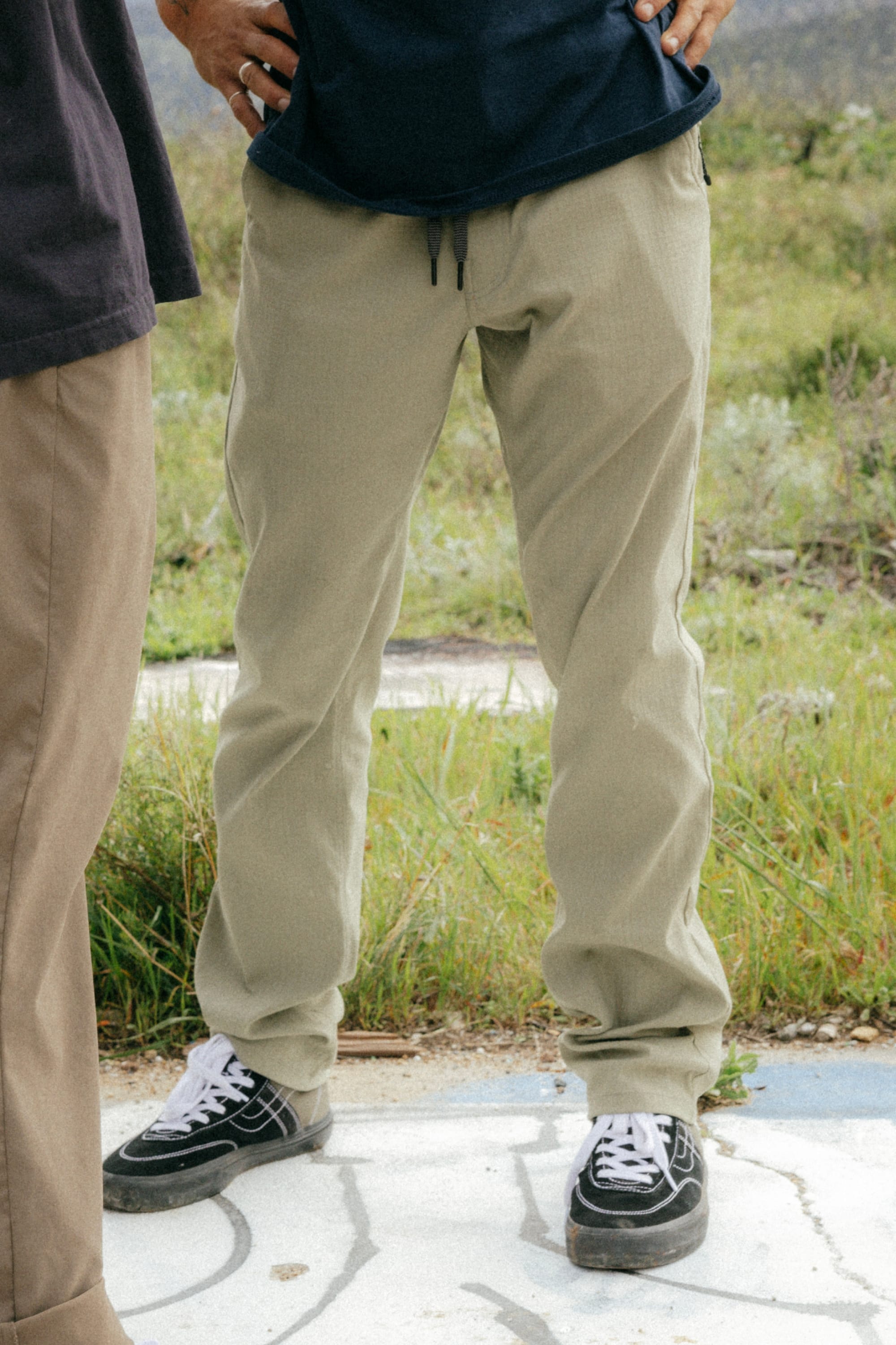 A person stands outdoors on concrete, hands on hips, wearing 686 Mens Everywhere Ripstop Pant - Slim Fit by 686 in light khaki and black sneakers. Grass and plants are in the background; another person is partially visible to their left.