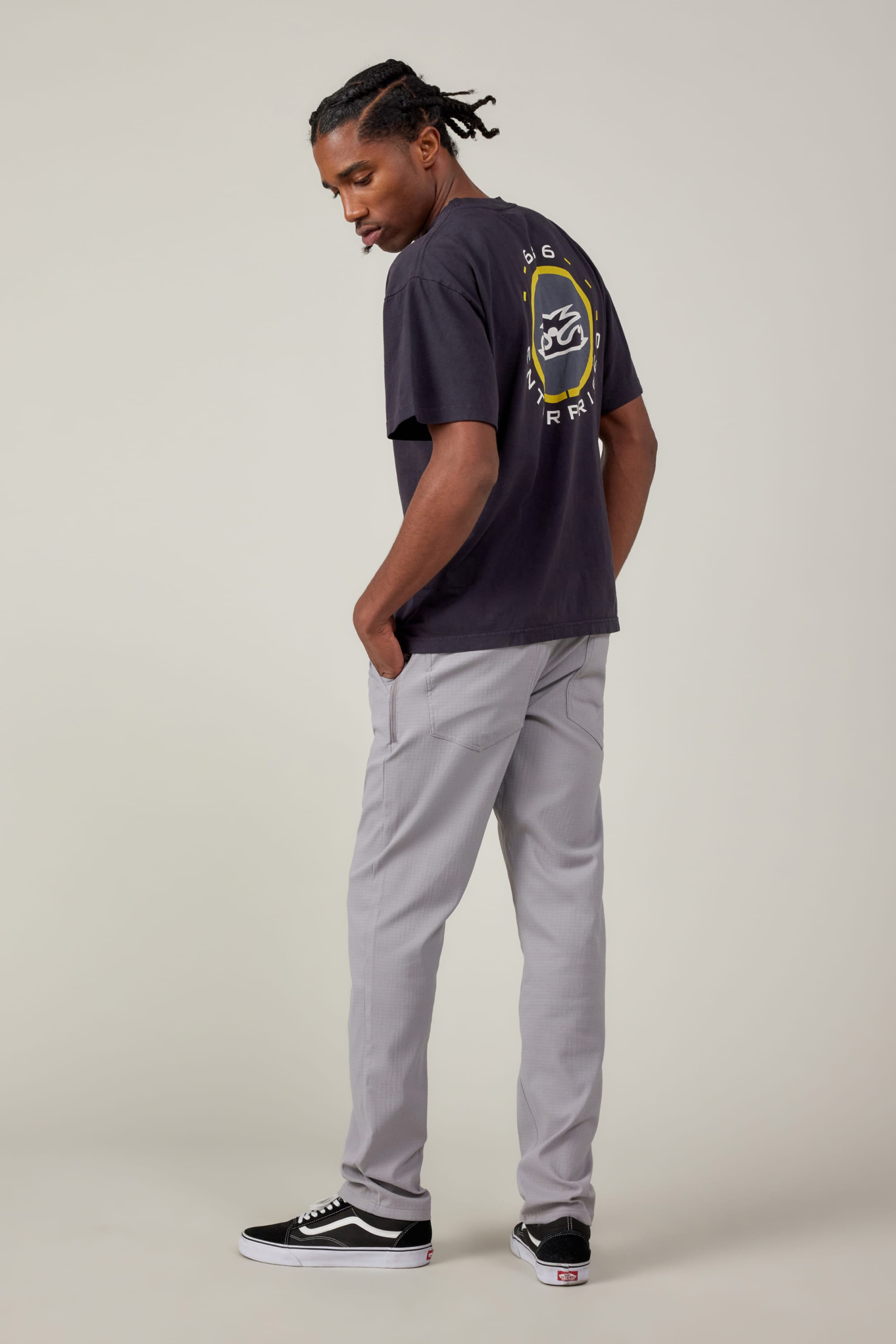 A person stands sideways with one hand in their pocket, wearing a black graphic t-shirt, 686 Mens Everywhere Ripstop Pant - Slim Fit by 686 in light grey, and black sneakers with white soles against a plain light background.