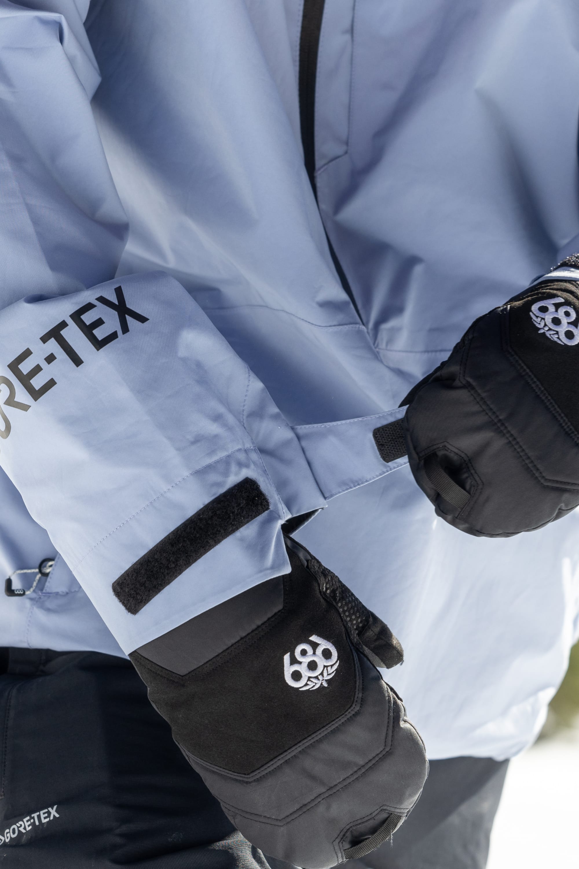 A person wearing black 686 gloves adjusts the velcro cuff of a light blue 686 Mens GORE-TEX Fragment Shell Anorak, highlighting the hands and jacket details for modern style on and off the mountain.