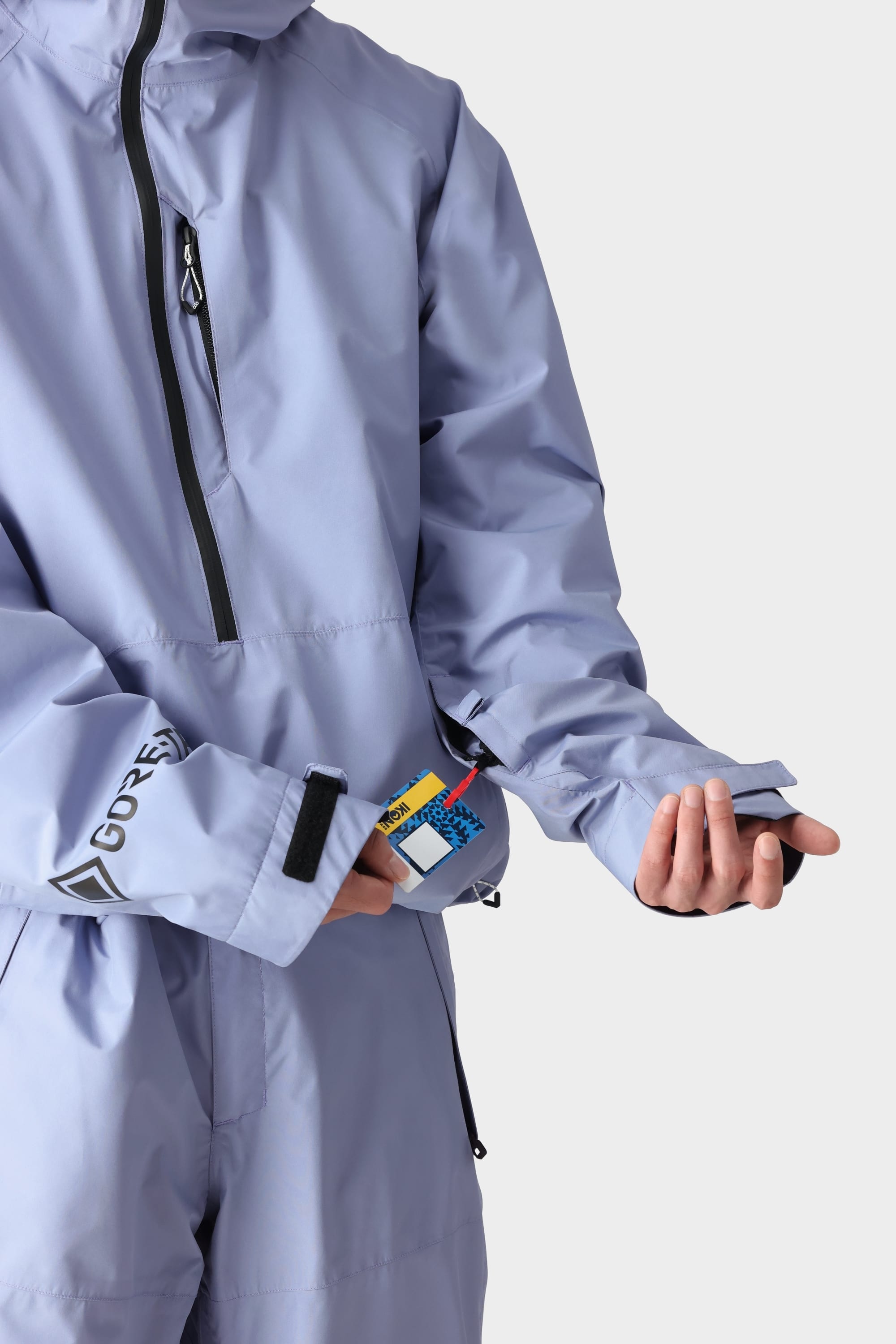 Wearing the 686 Mens GORE-TEX Fragment Shell Anorak, a person pulls ski passes and a card from a small zippered sleeve pocket on their left arm—ready for adventures. Only their torso and arms are visible against a plain background.