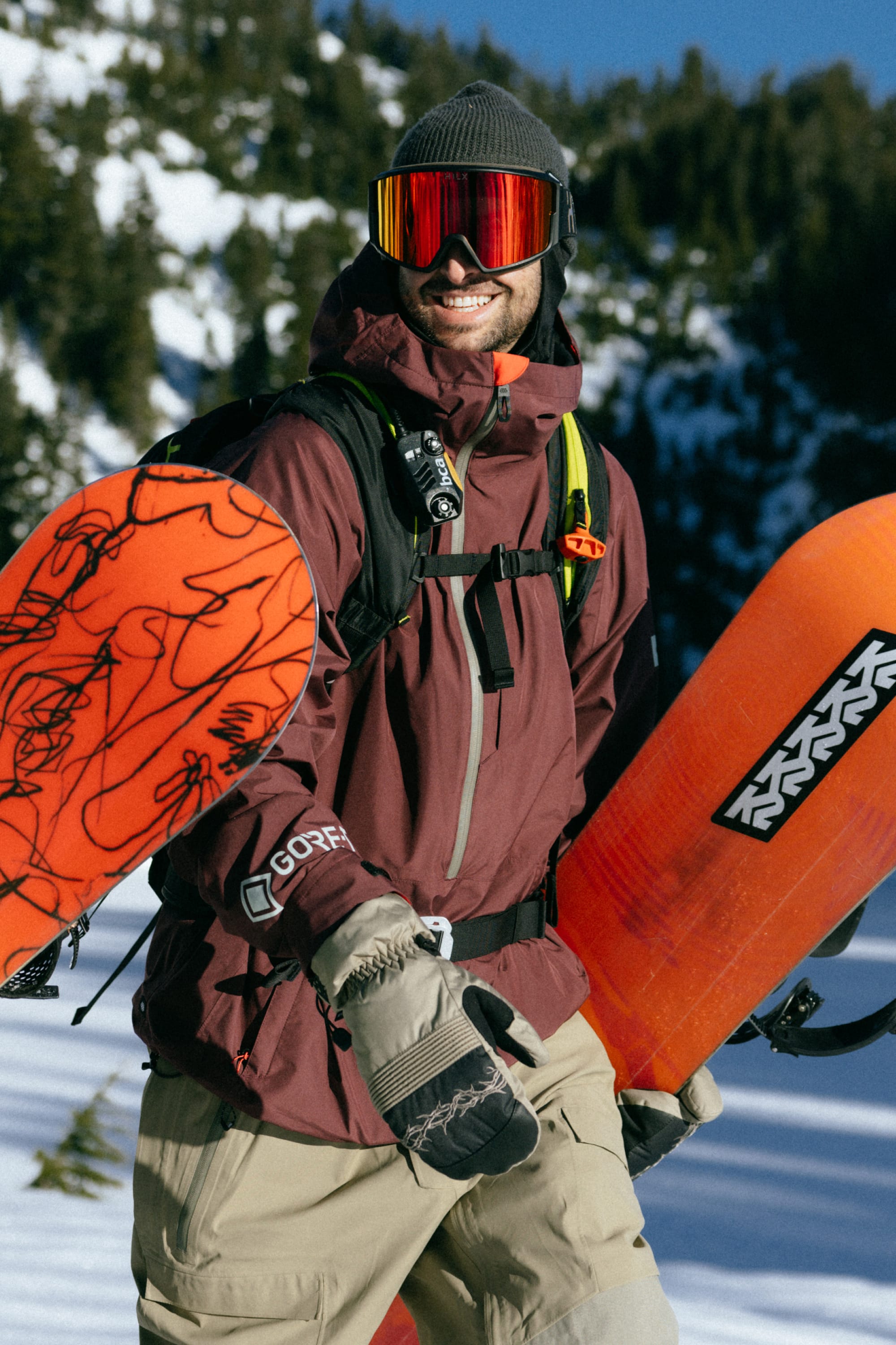 A snowboarder wearing the 686 Mens GORE-TEX Fragment Shell Anorak, beige pants, and reflective goggles stands in the snow holding two orange snowboards. Pine trees and snowy slopes frame a scene ready for adventure.