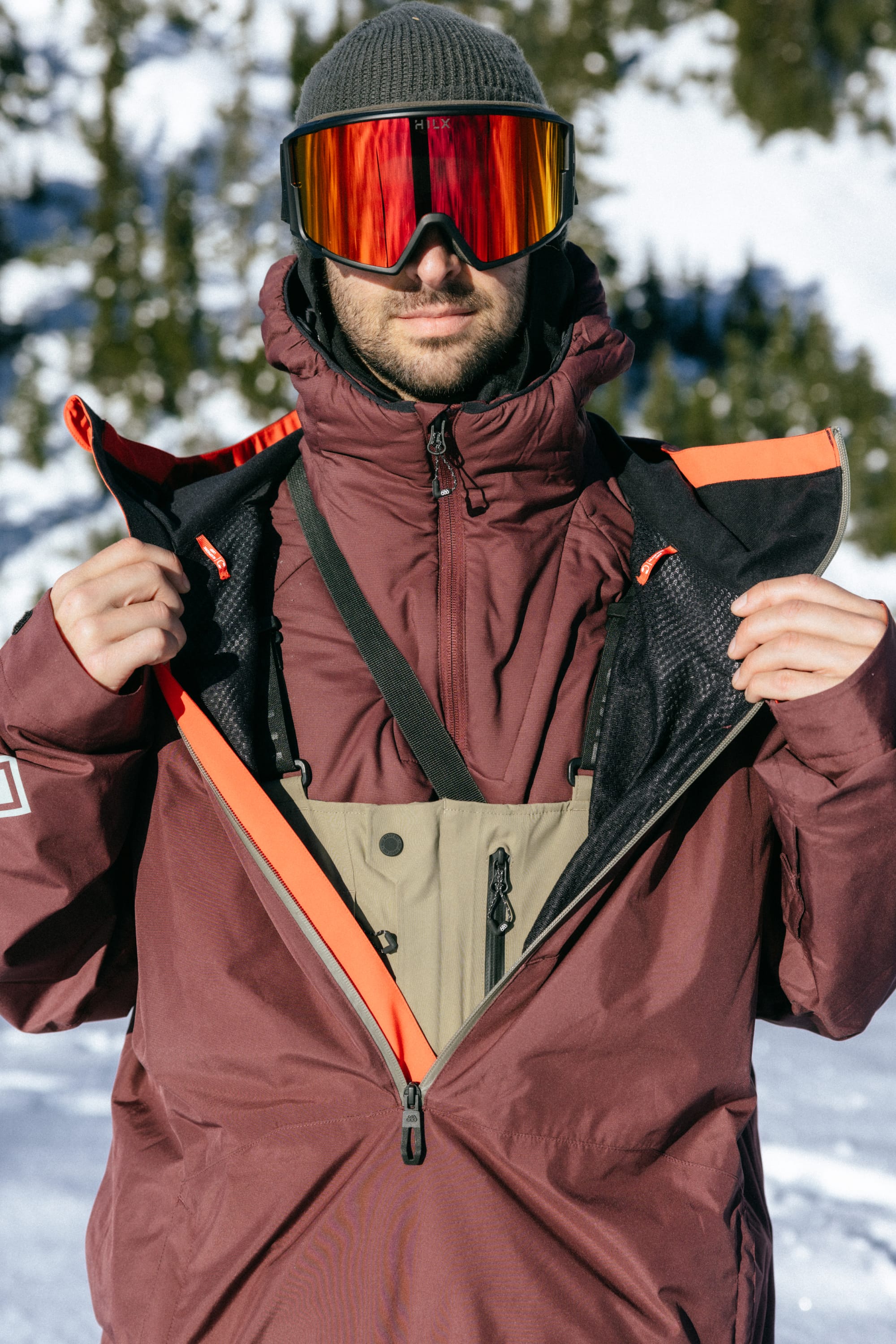 Wearing a 686 Mens GORE-TEX Fragment Shell Anorak, a gray beanie, and reflective ski goggles, a person stands in snowy outdoors unzipping their jacket to reveal an inner layer, with snow-covered trees in the background.