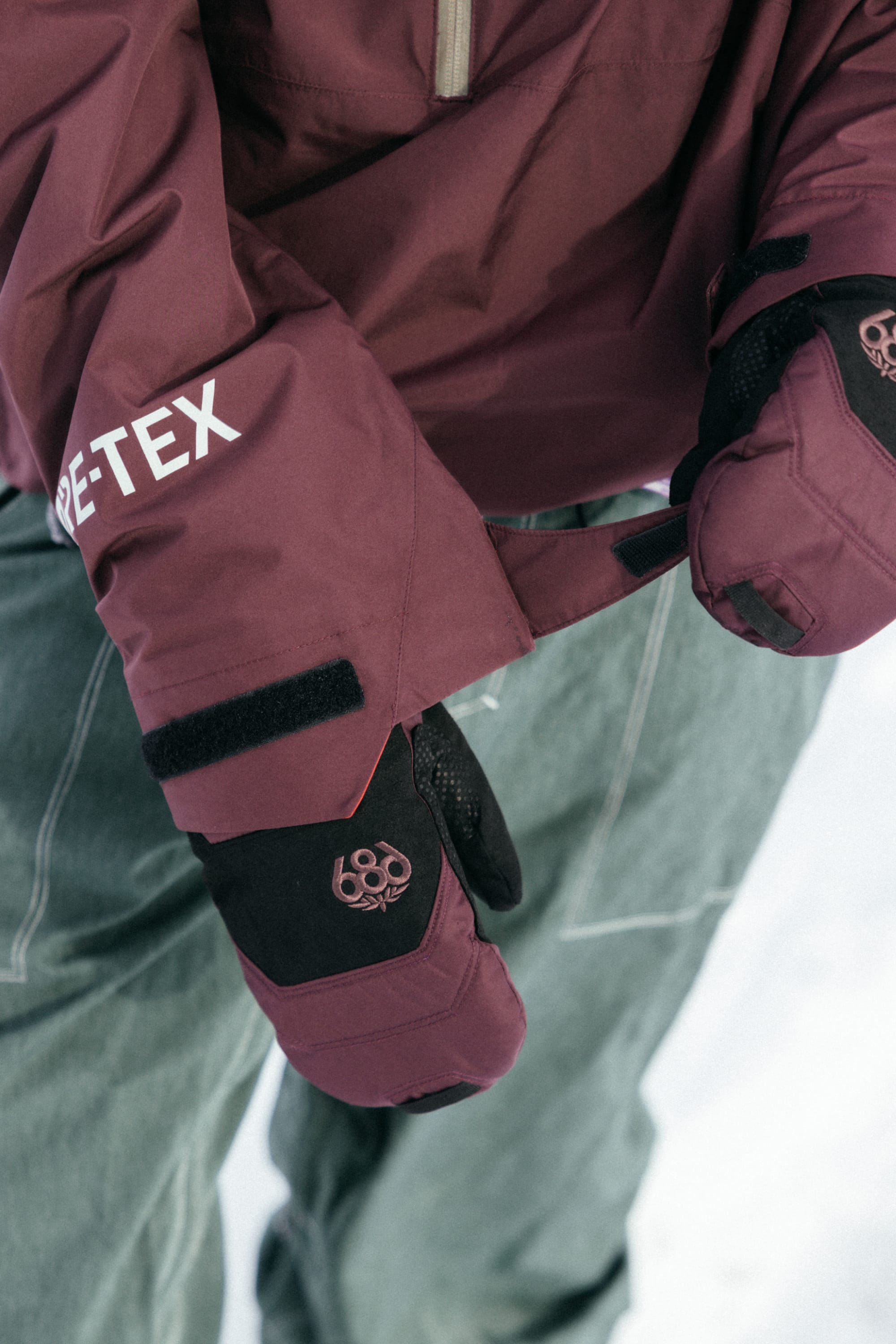 A person in the 686 Mens GORE-TEX Fragment Shell Anorak, black winter gloves, and green pants adjusts their cuff on snow—highlighting this modern 686 jacket designed for comfort on and off the mountain.
