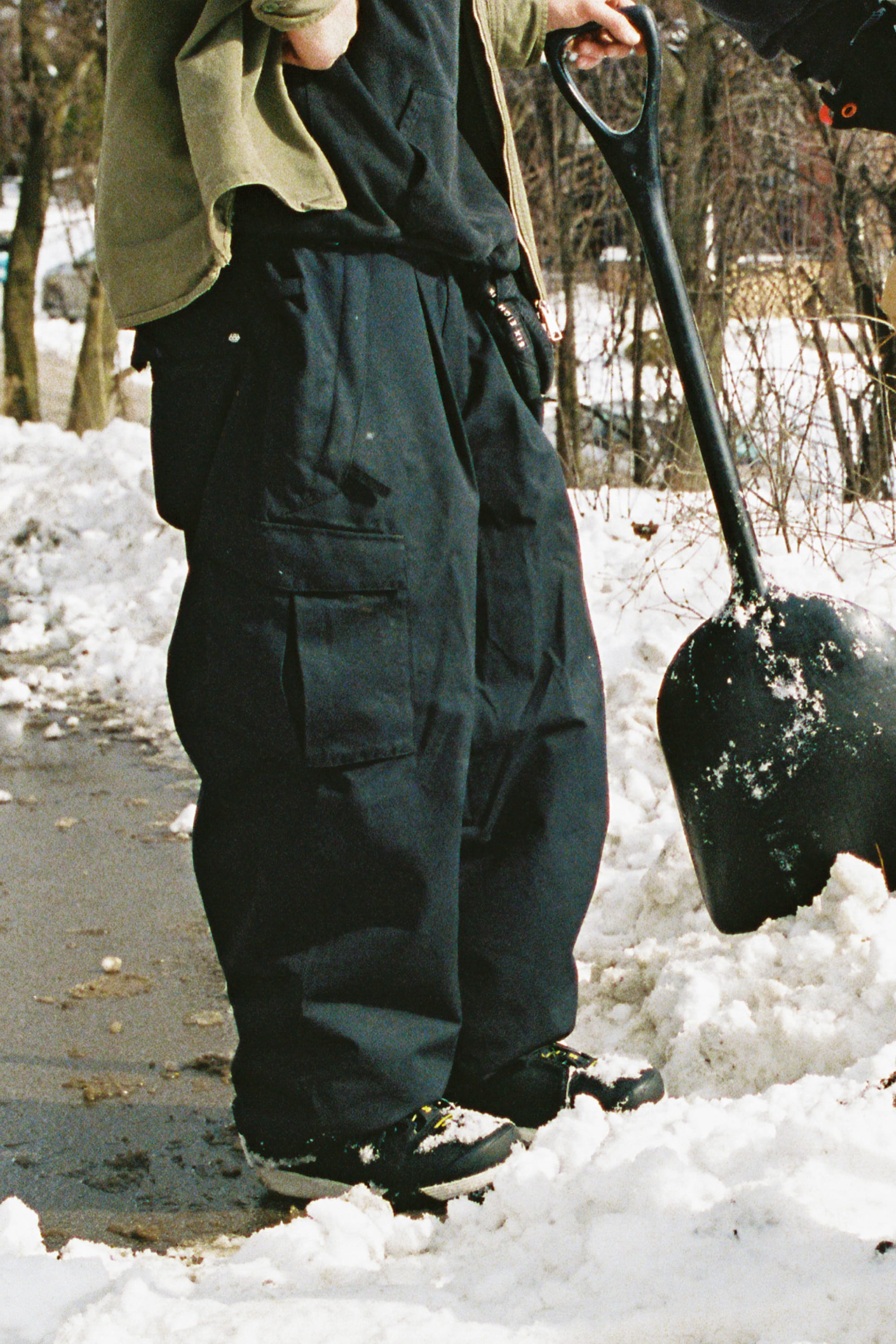 Wearing the 686 Men’s Essox™ Cargo Pant, a person stands on a snowy sidewalk holding a black snow shovel, with snow and trees in the background.